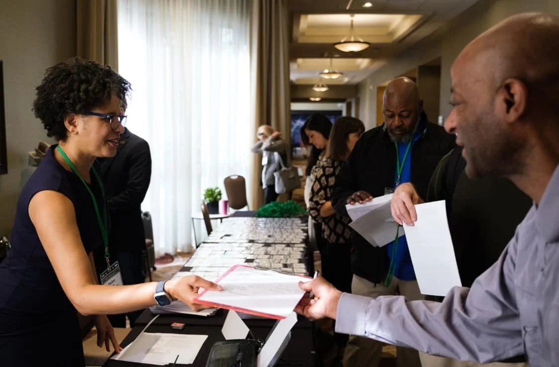 A woman with glasses and short curly hair is handing a document to a man with a shaved head. They are at a conference or event registration table, smiling and engaging in conversation. Several people are in the background, in a room with curtains and