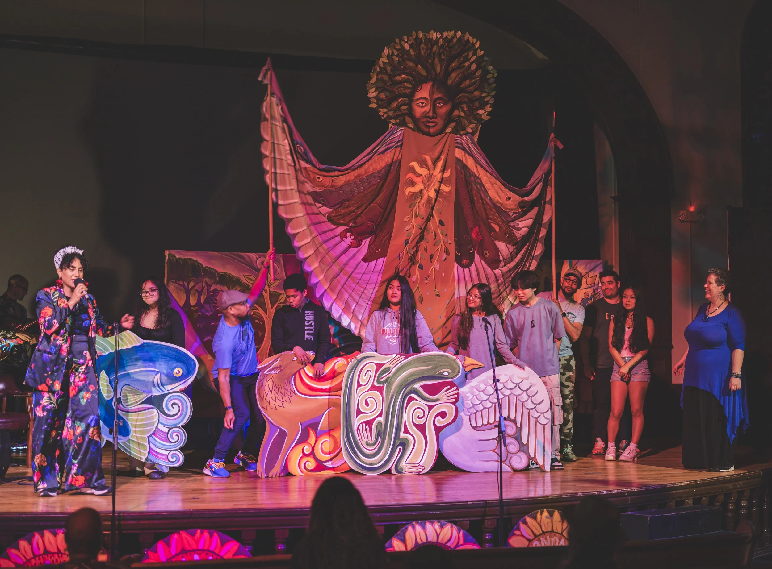 Group of people on stage during a performance or event, with large colorful artistic figures and a backdrop featuring a butterfly and other vibrant designs.