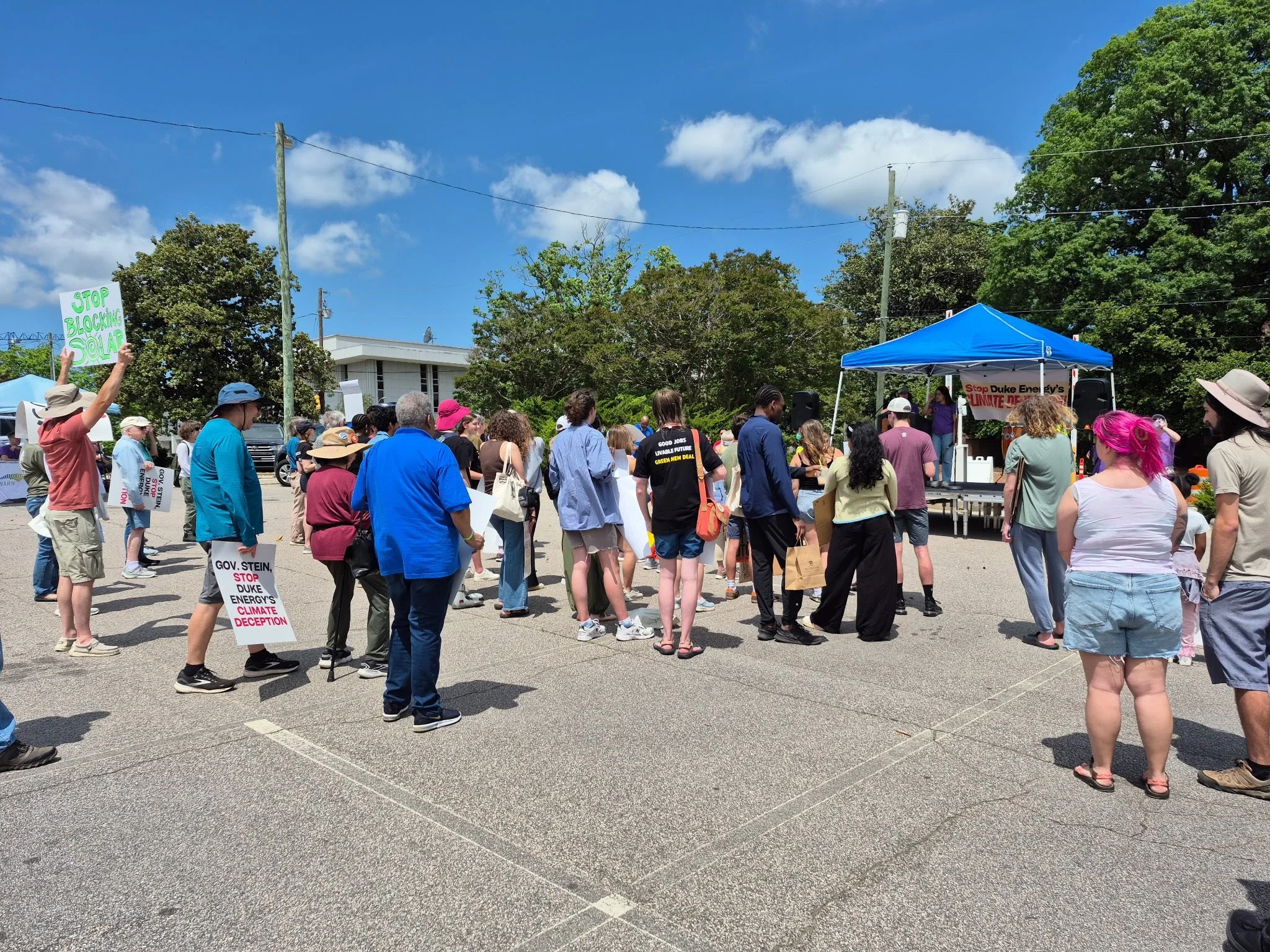 A diverse group of people protesting outdoors on a sunny day, holding signs calling for action on climate change, standing in front of a stage with a speaker and a blue canopy.