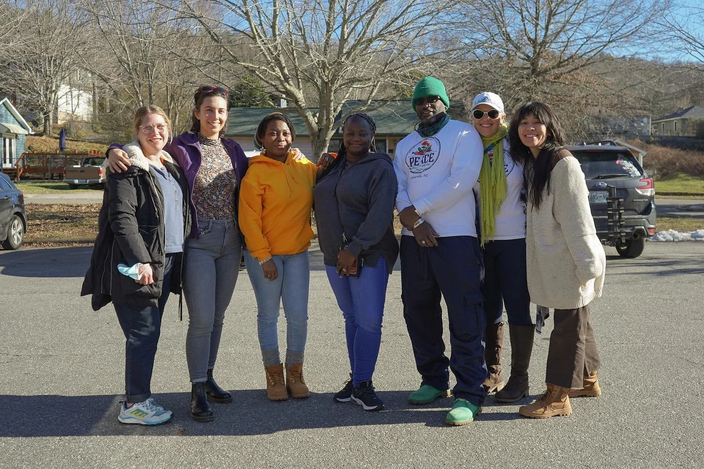 Group of diverse people standing outdoors in a suburban neighborhood, smiling at the camera, with leafless trees and parked cars in the background.