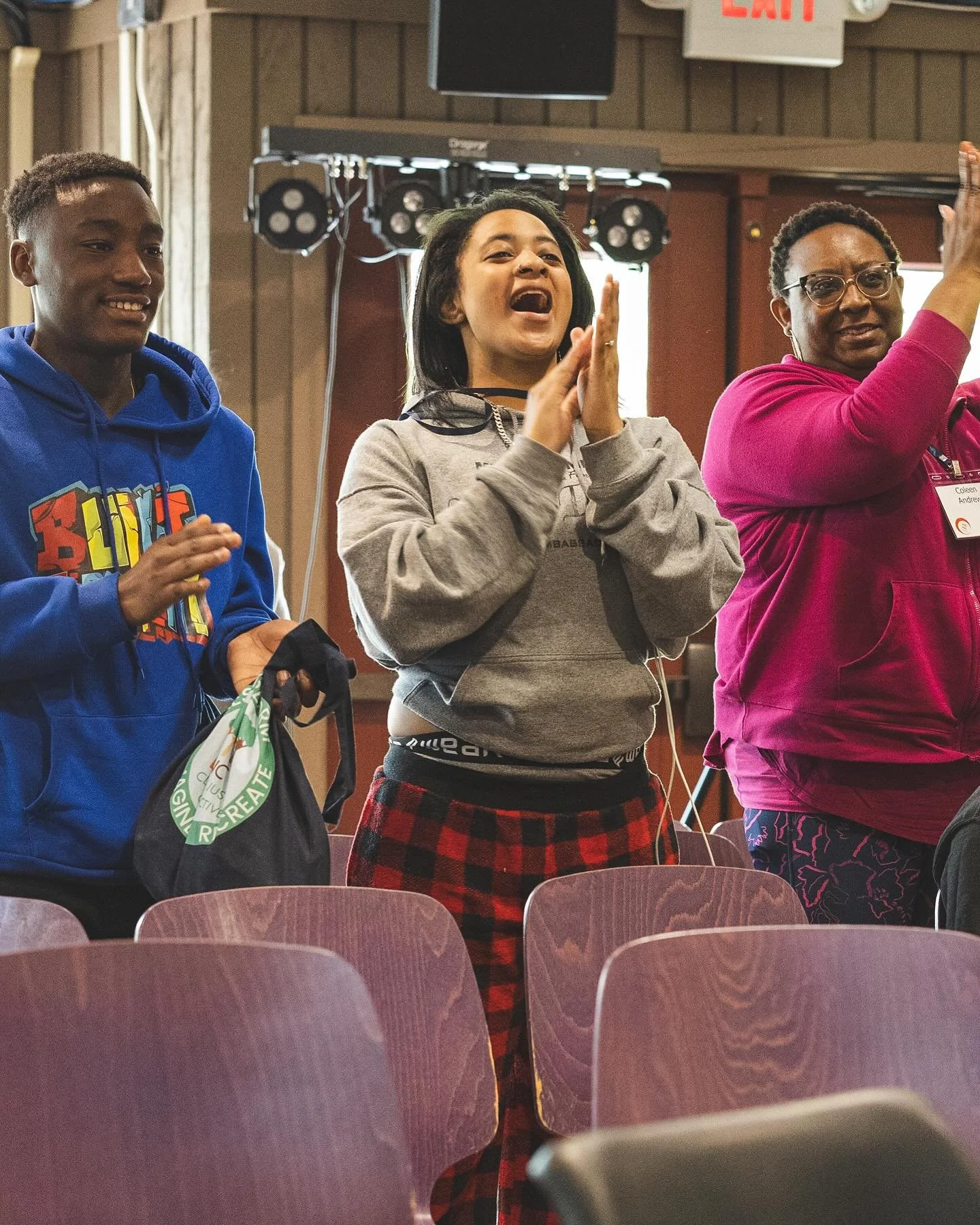 Three people standing and smiling at an indoor event, with chairs in the foreground and stage lighting in the background.