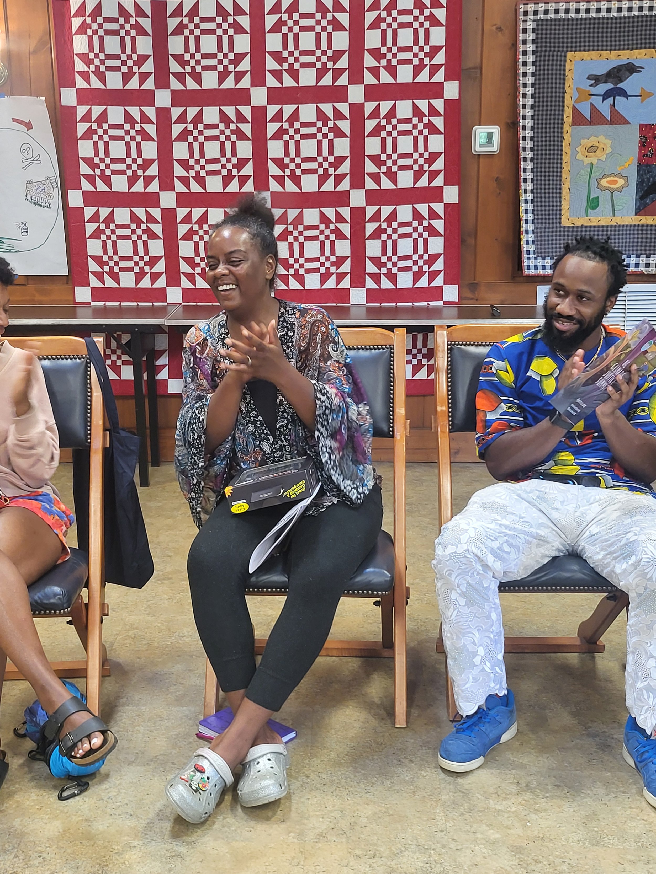 Three people sitting in a row, smiling and laughing, in a room with colorful wall hangings and wooden walls.