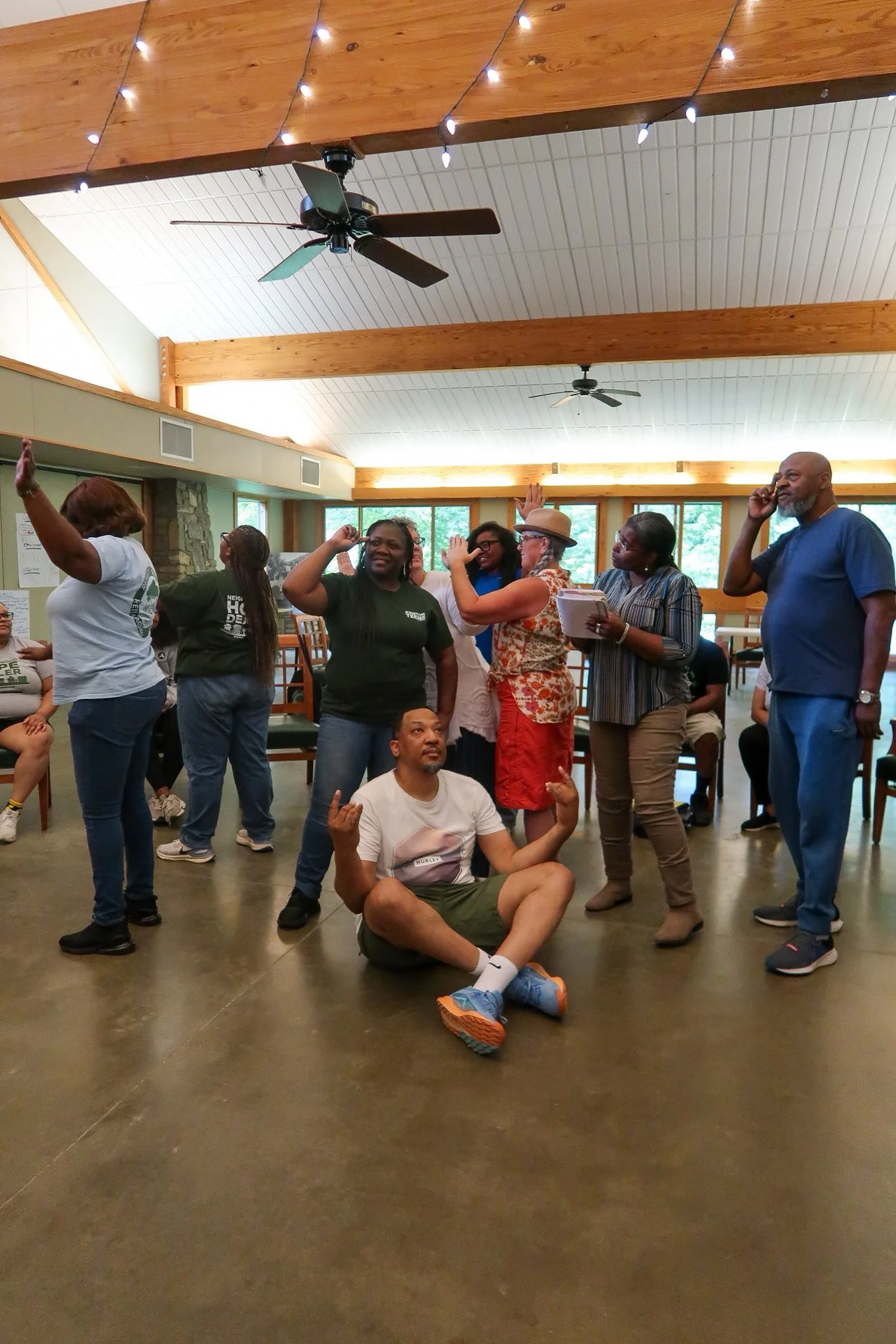 Group of people enjoying a social gathering indoors, some standing and one person sitting on the floor, with string lights and wooden beams on the ceiling.