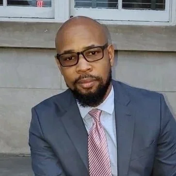 A man with glasses and a beard in a suit, sitting outside in front of steps, looking at the camera.