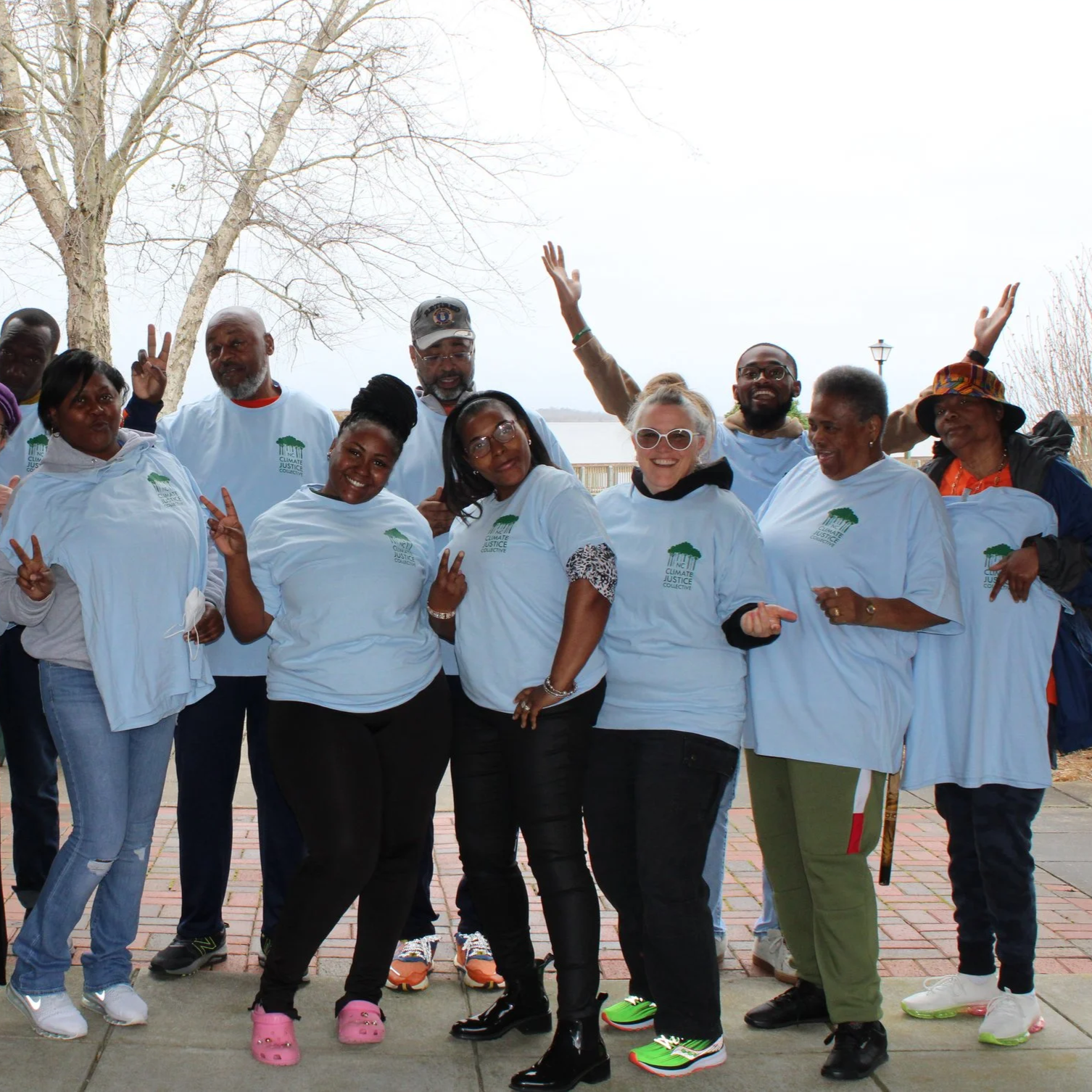 Group of diverse people outdoors wearing matching white T-shirts with a green tree logo, smiling and posing for a photo during daylight.