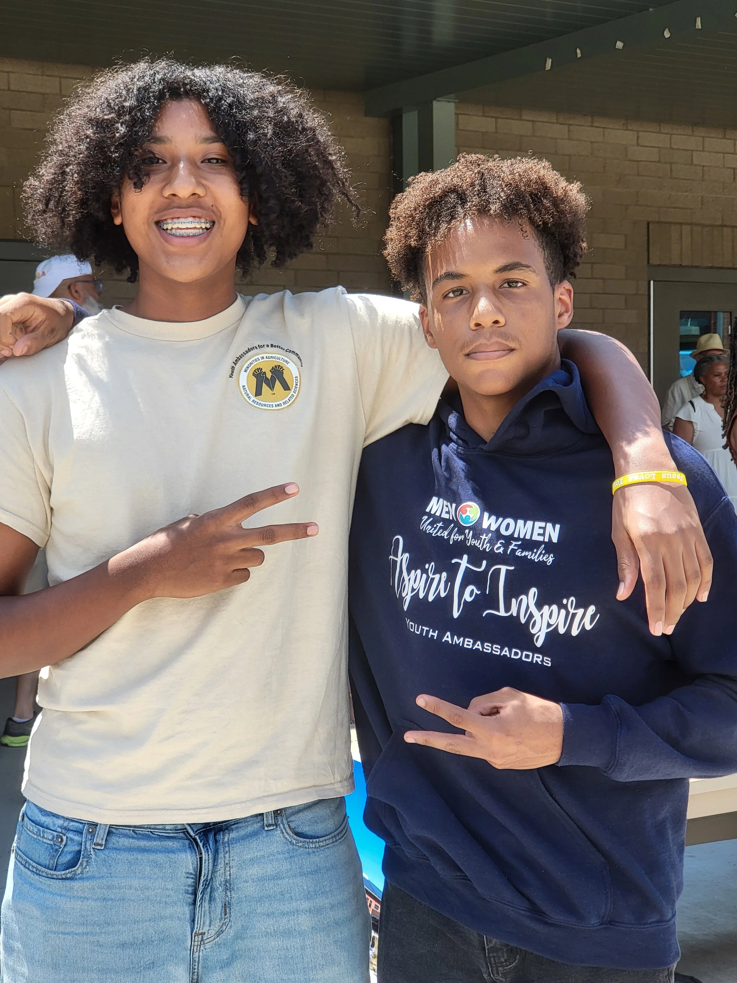 Two young people standing together outdoors, both making peace signs with their hands. The person on the left wears a light-colored T-shirt with a circular logo, has curly dark hair, and braces. The person on the right wears a dark hoodie with white 