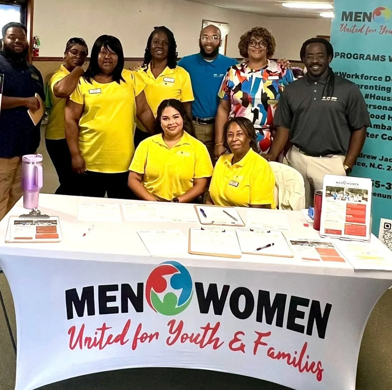 Group of men and women standing behind a table with a 'MEN WOMEN United for Youth & Families' banner, at an indoor event or conference.