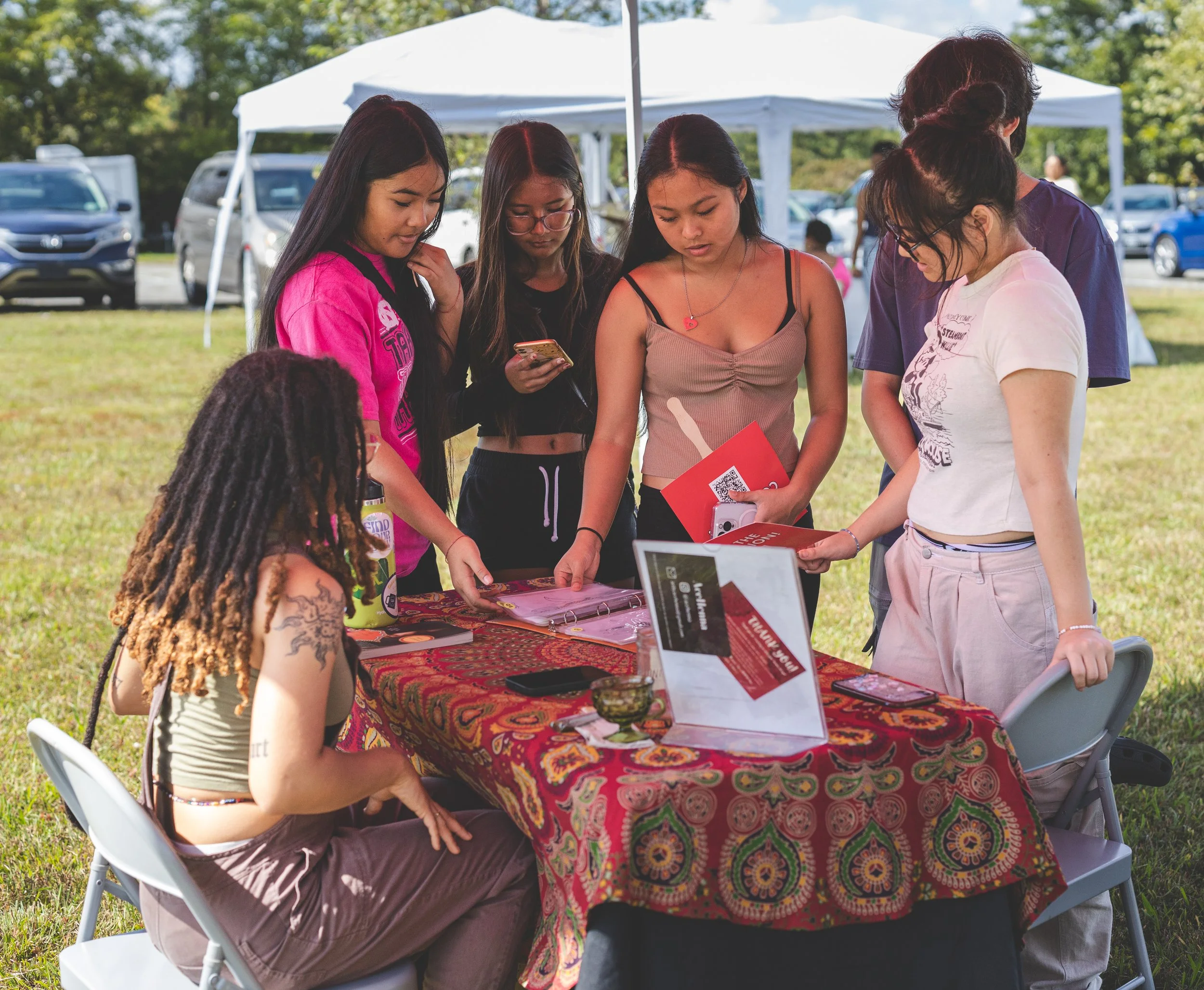 A group of seven young women gathered around a table outdoors, looking at various items and documents, with a white canopy tent and parked cars in the background.