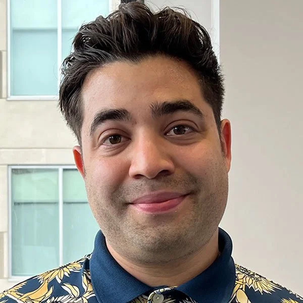 Close-up of a man with dark hair, wearing a patterned shirt, smiling in front of a building with large windows.