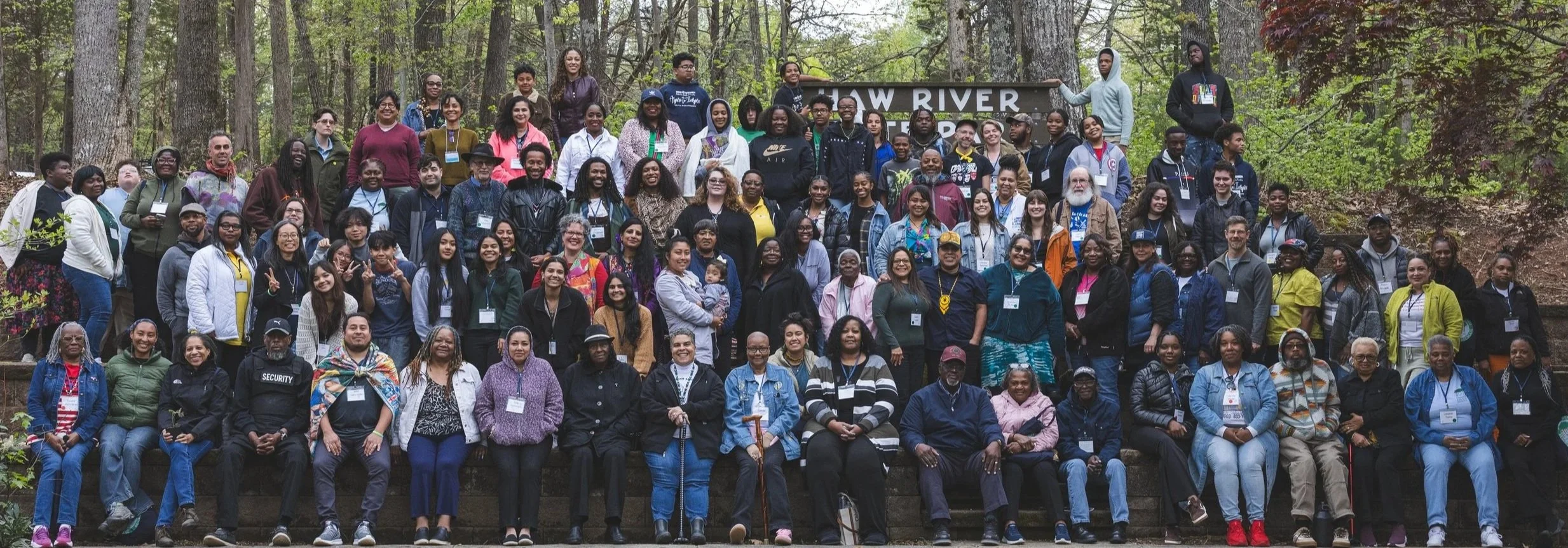 Large group photo of an intergenerational multiracial coalition meeting in a forest, standing on outdoor steps, with a sign that reads 'LOW RIVER' in the background.