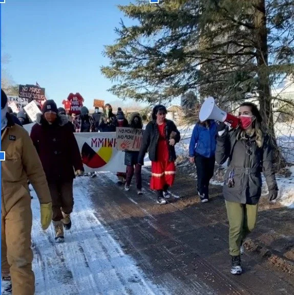 Group of people participating in a protest march outdoors on a snowy day, some holding signs and a person speaking into a megaphone.