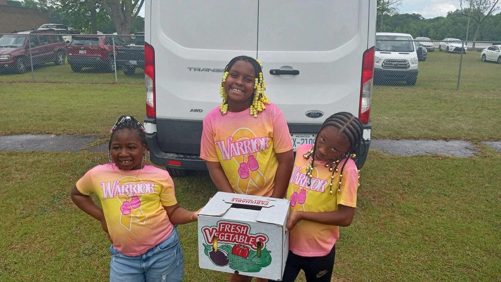 Three young girls standing on grass holding a box of fresh vegetables, smiling, wearing matching pink and yellow T-shirts with boxing gloves and the word 'WARRIOR' printed on them.