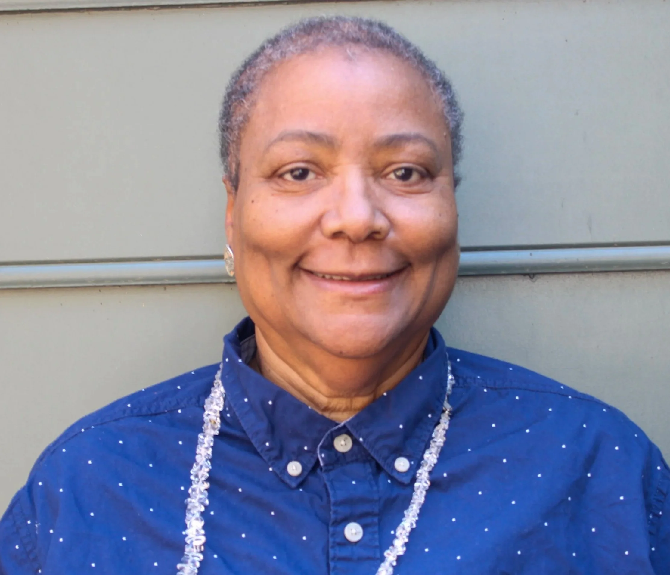 A smiling woman with short gray hair, wearing a blue button-up shirt with white specks, a glittery necklace, and earrings, standing in front of a gray wall.