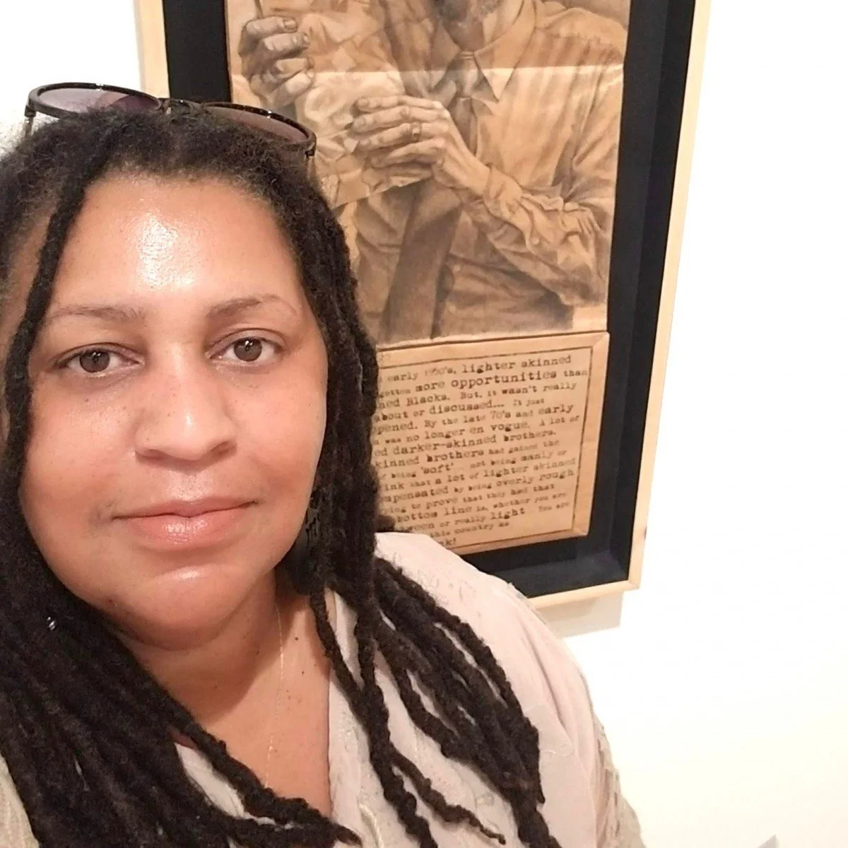 A woman with dreadlocks and sunglasses on her head posing for a selfie in front of a framed artwork featuring a person in a beige shirt with a ribbon tied at the neck and a section of text.