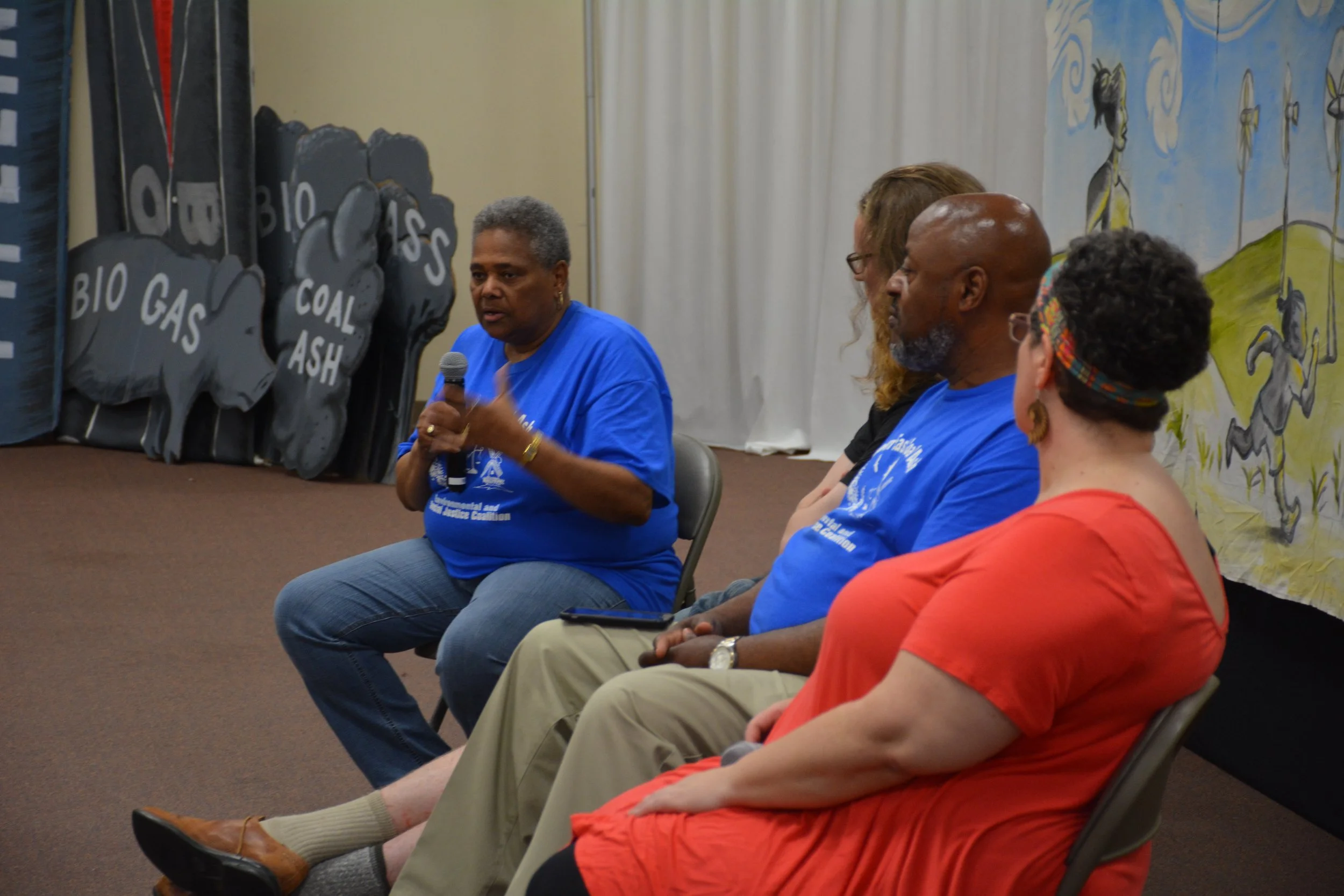 A woman in a blue shirt speaking into a microphone while seated among three other people, with a backdrop of a mural featuring a woman, wind turbines, and horses.