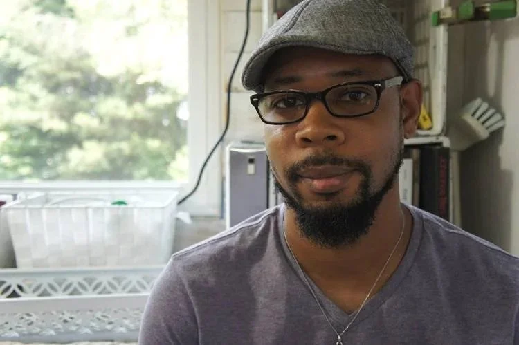 A man wearing a gray cap, glasses, and a gray t-shirt sitting indoors with a background of a window, a white plastic container, a bookshelf, and some books.