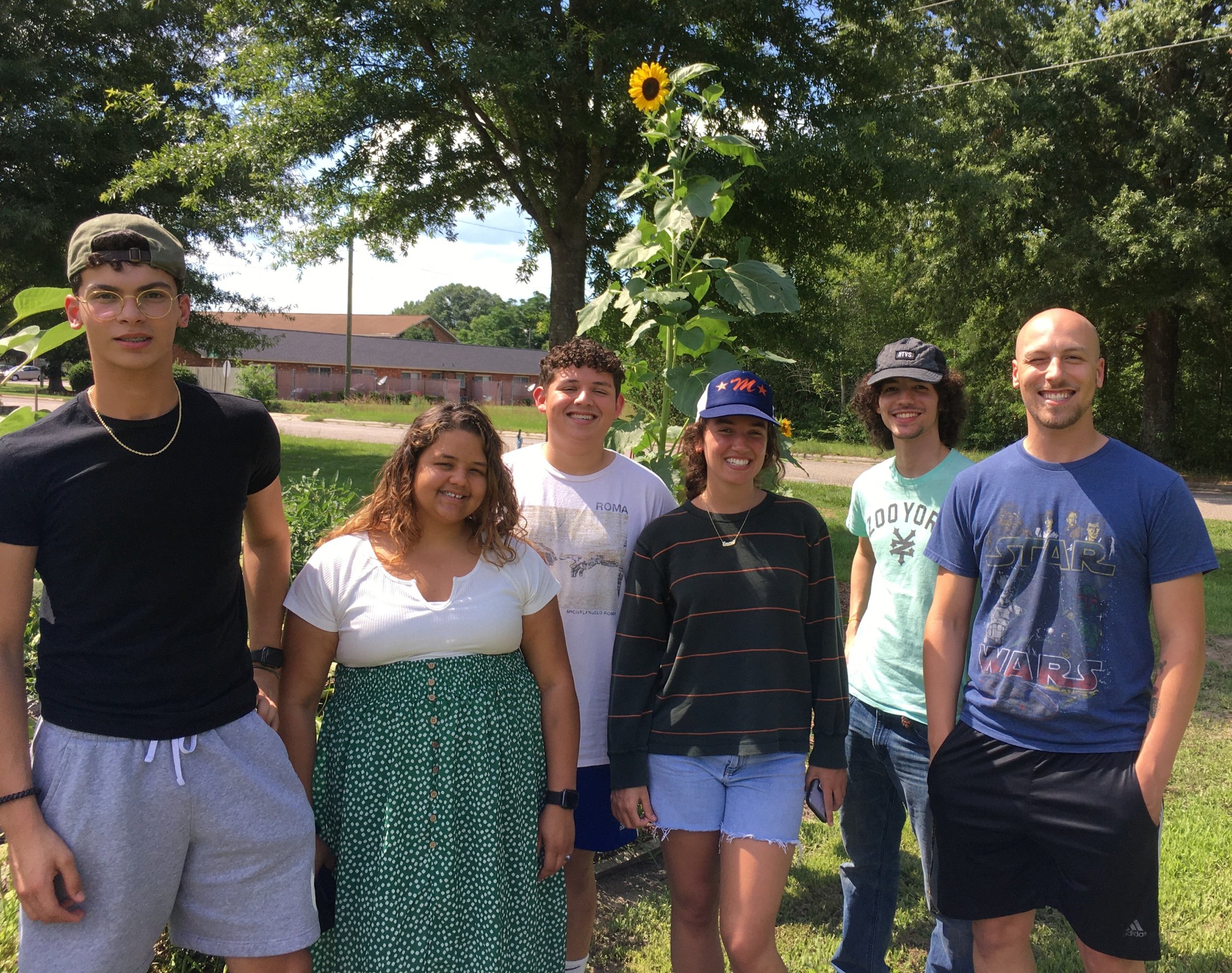 Group of seven young adults smiling outdoors, standing in front of a sunflower plant with a large tree and a building in the background.