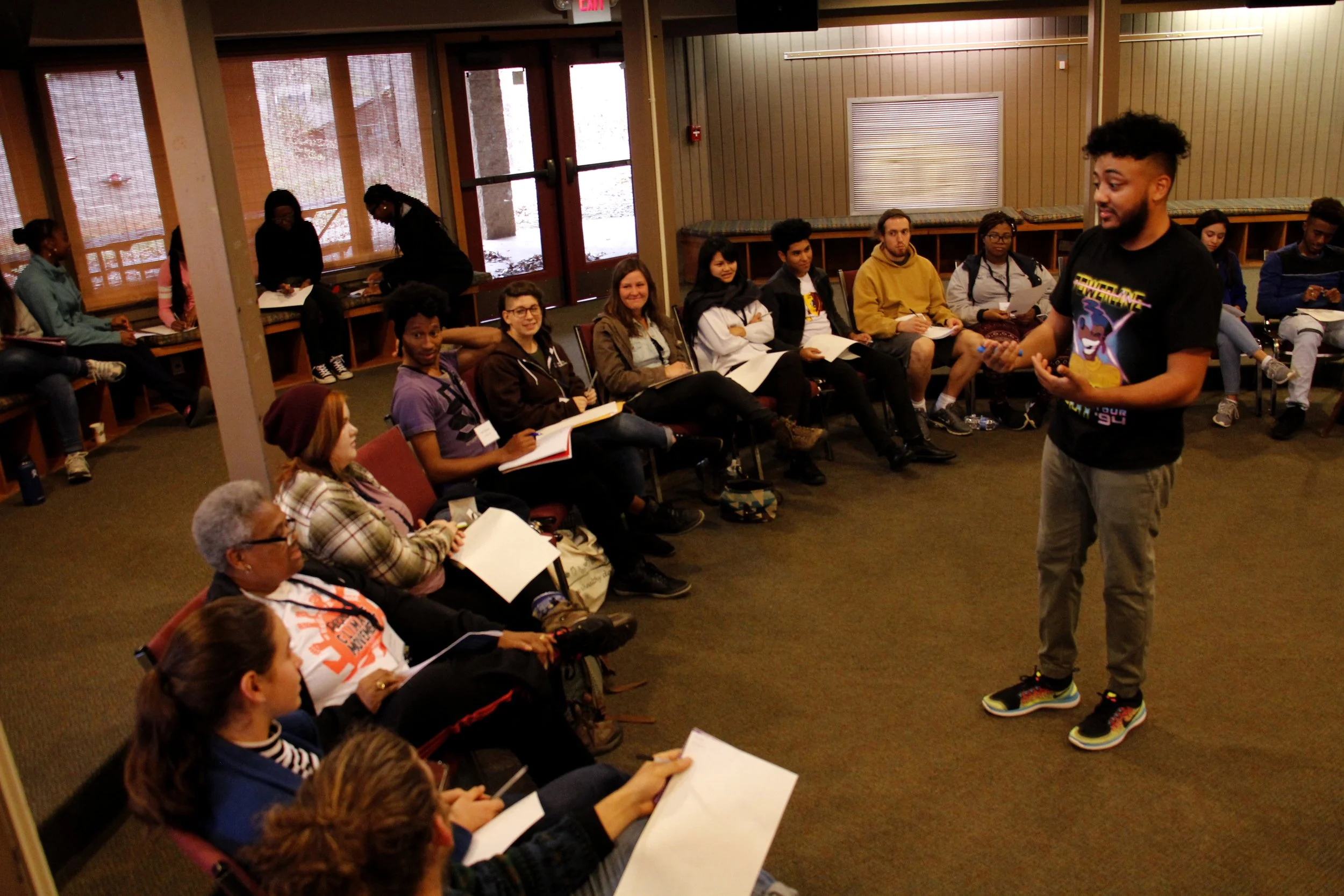 A young man with an afro hairstyle and casual clothing giving a presentation or speech in front of a seated audience in a conference room with large windows.