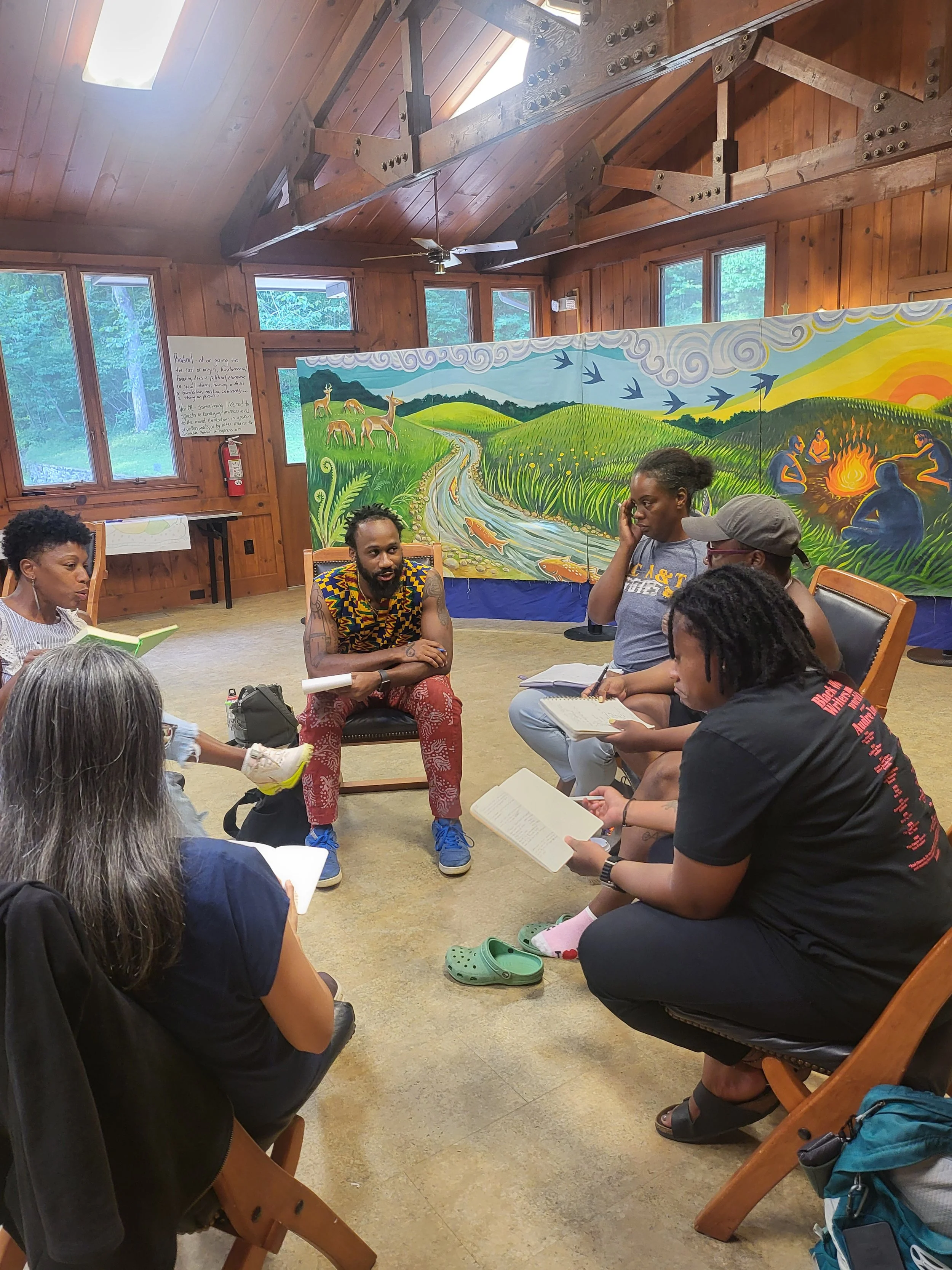 A group of people seated in a circle in a wooden room, participating in a discussion or meeting, with a colorful mural of a nature scene in the background.