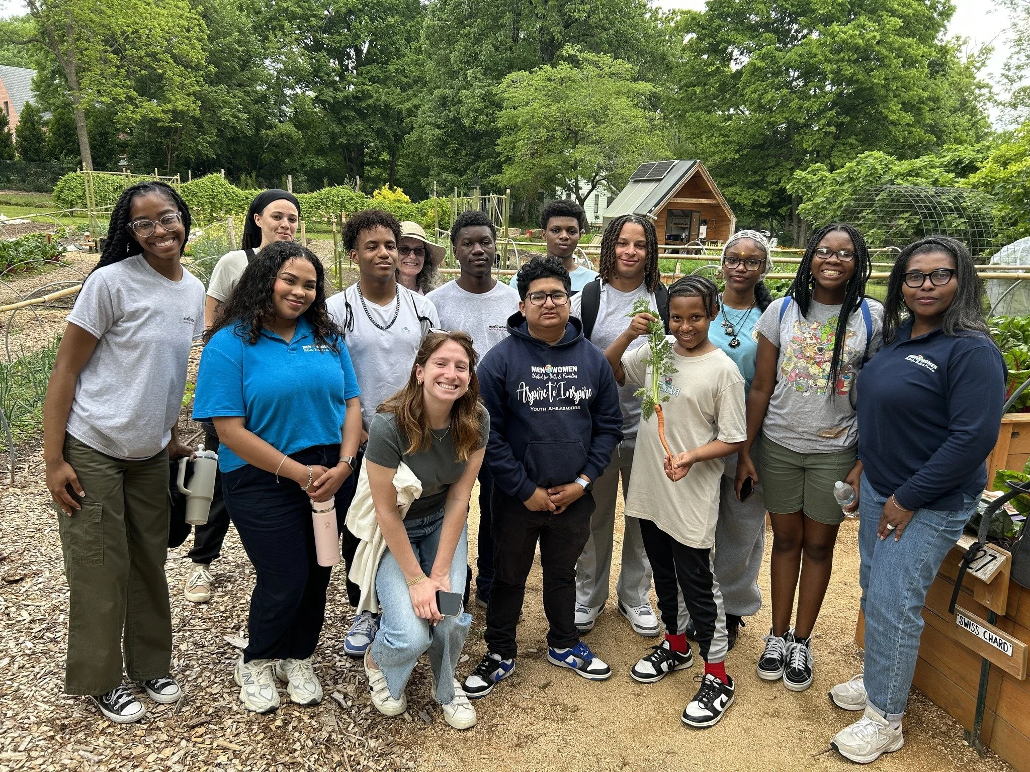 Group of young people and adults standing in a community garden, with trees and garden beds in the background.