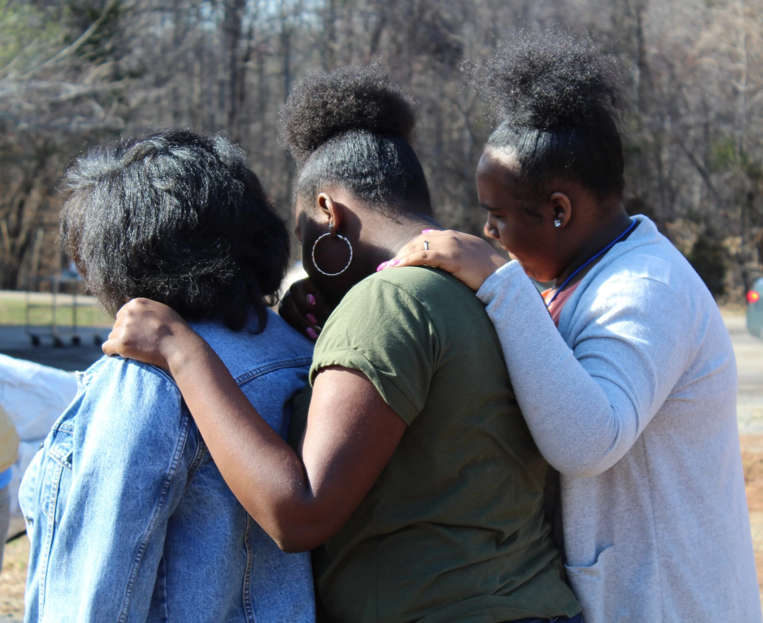 Three women standing closely together, hugging with their heads bowed and eyes closed, outdoors on a sunny day.