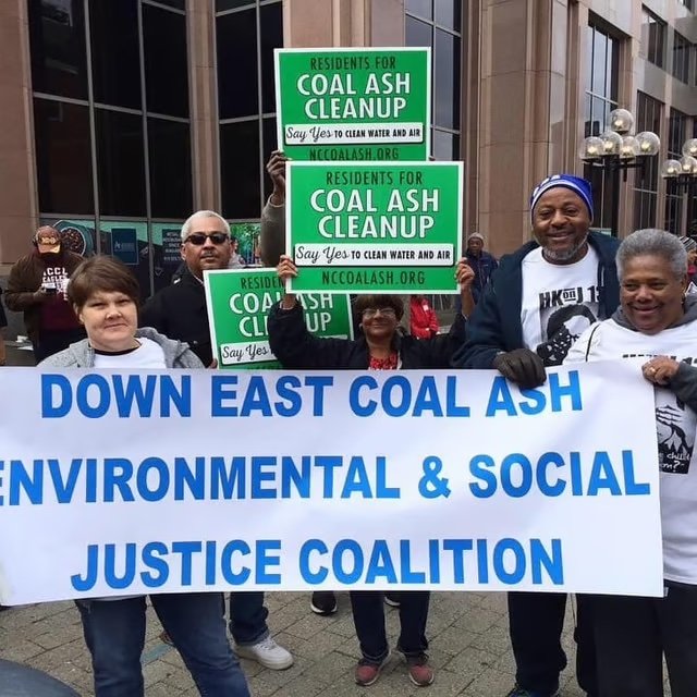 Group of people holding signs and a banner protesting coal ash cleanup and environmental justice in downtown East with buildings in the background.