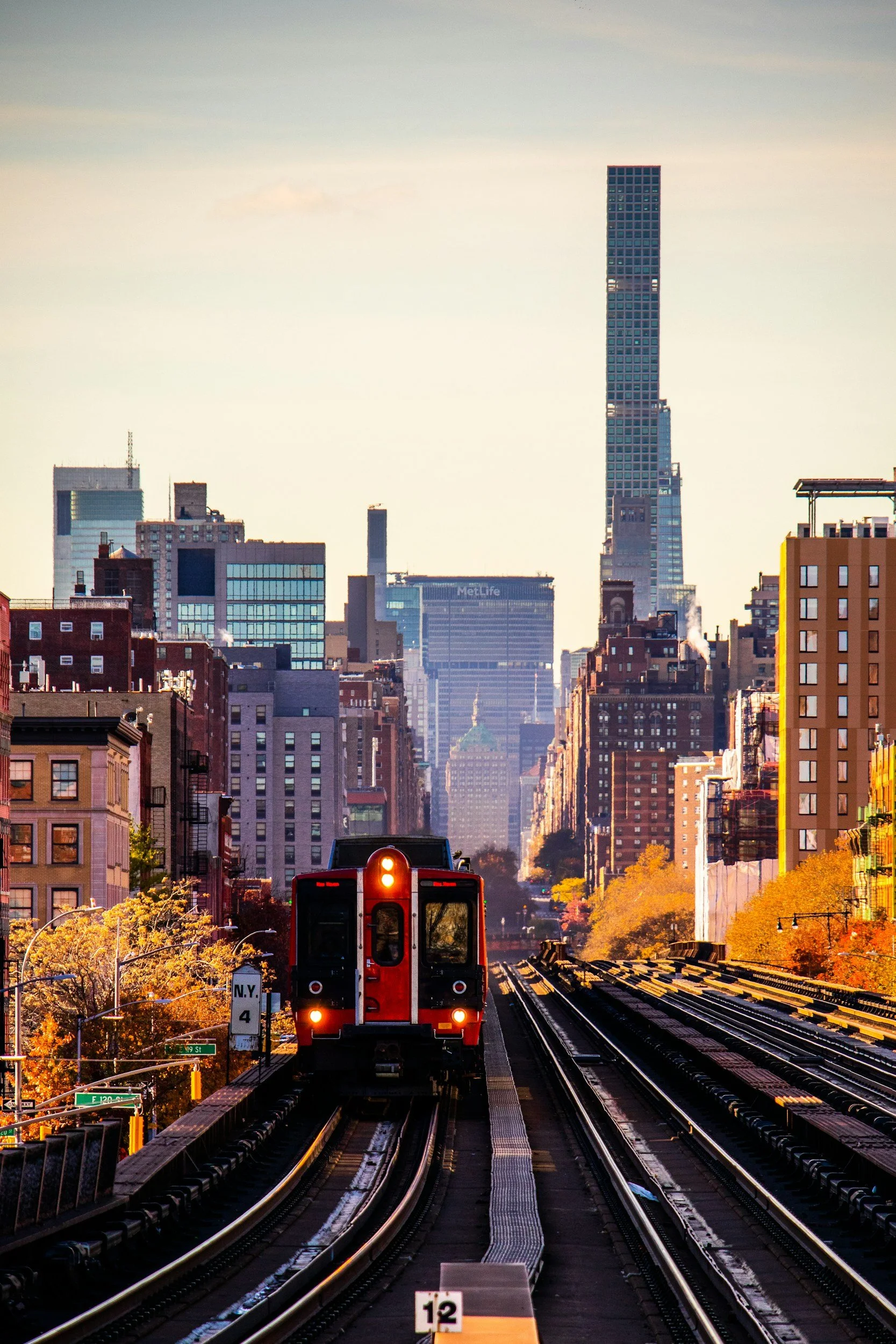 Red train on elevated tracks in a city with tall buildings, including a skyscraper, during sunset with orange and yellow trees along the sides.
