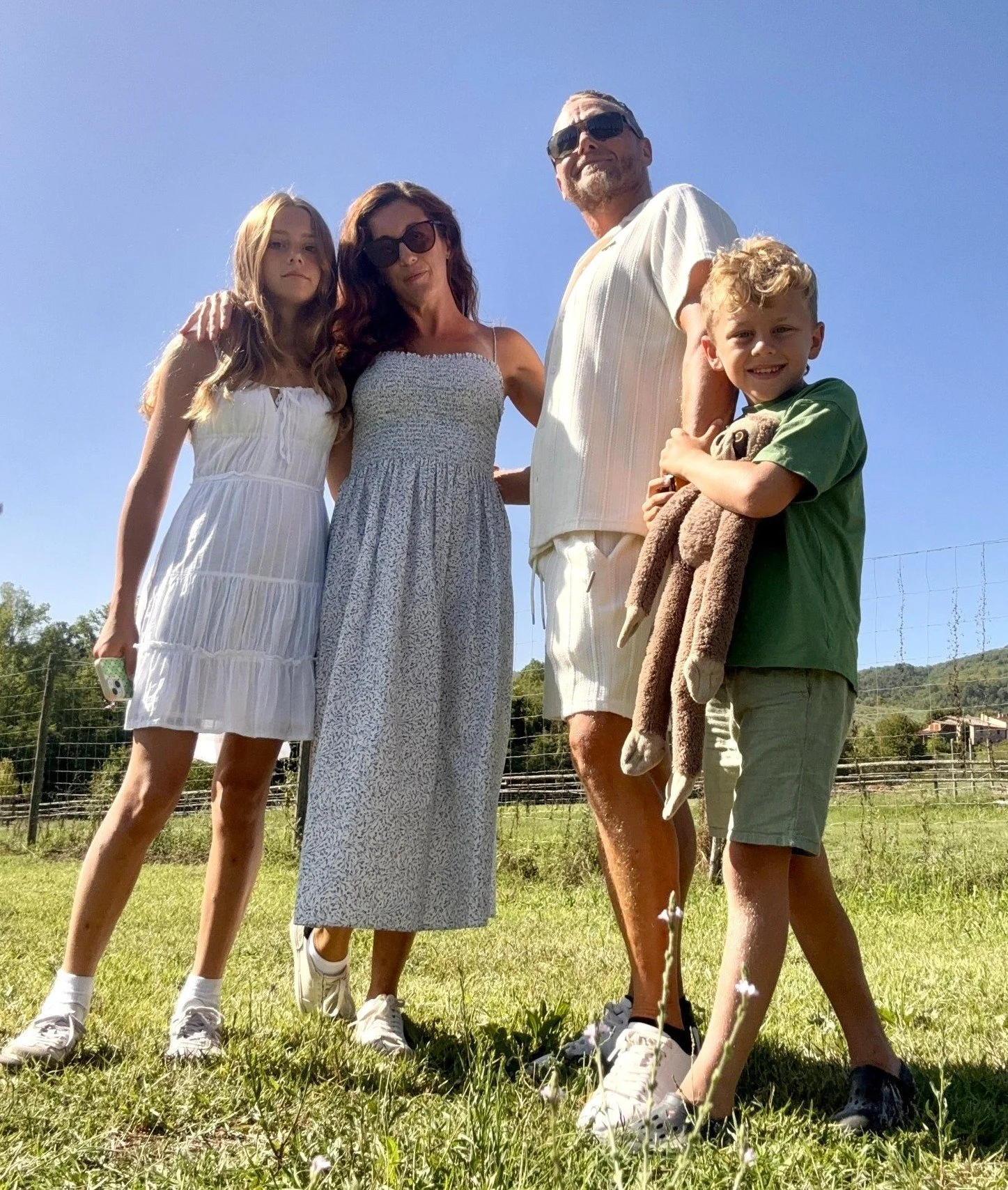 A family of five standing outdoors on grass under a clear blue sky, with a fence and trees in the background. The family includes a woman, a man, two girls, and a boy holding a stuffed animal.