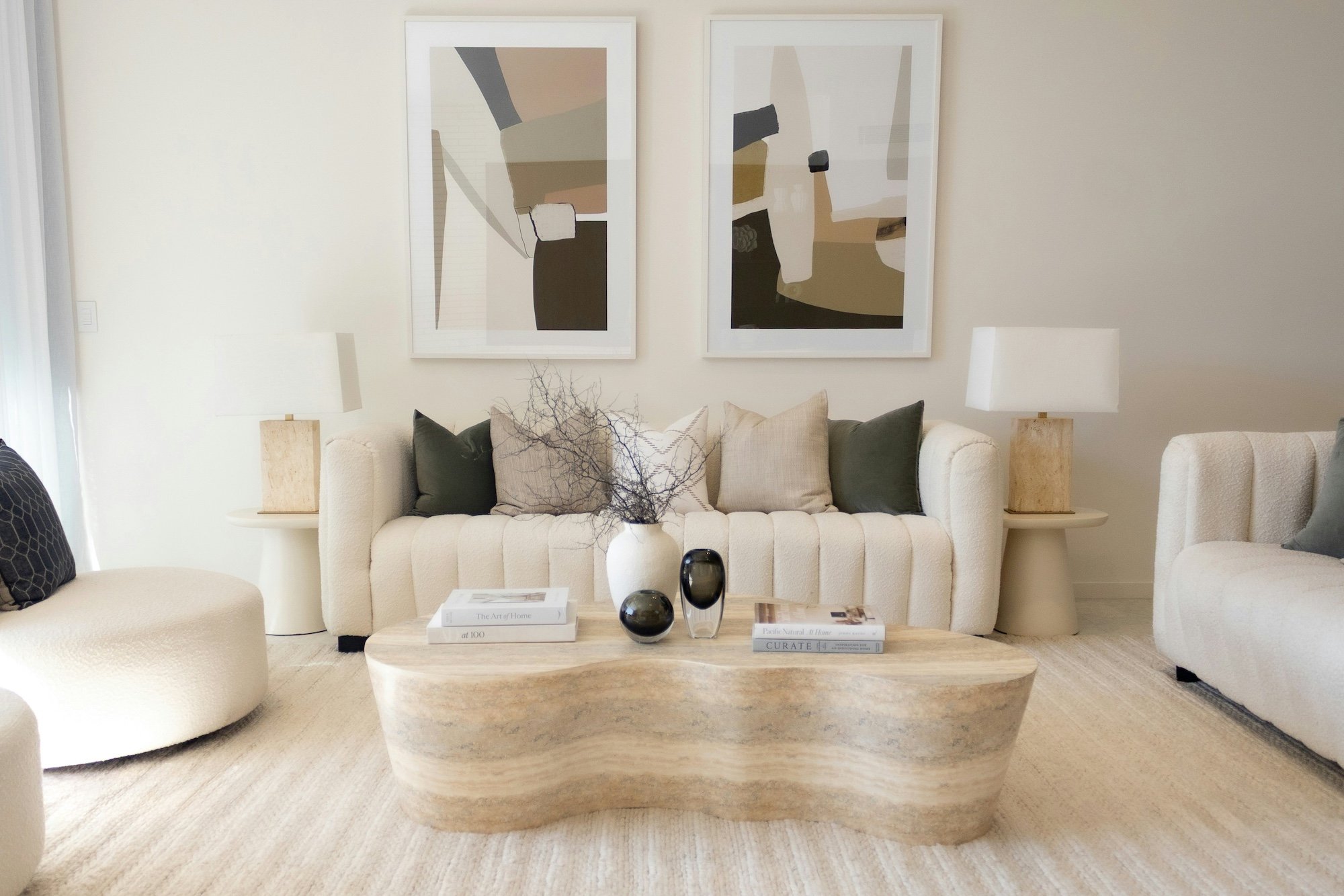 A contemporary living room with two abstract paintings, two white lamps, and a curved beige sofa with assorted pillows. There is a wooden coffee table with books and decorative vases, and a light-colored textured rug on the floor.