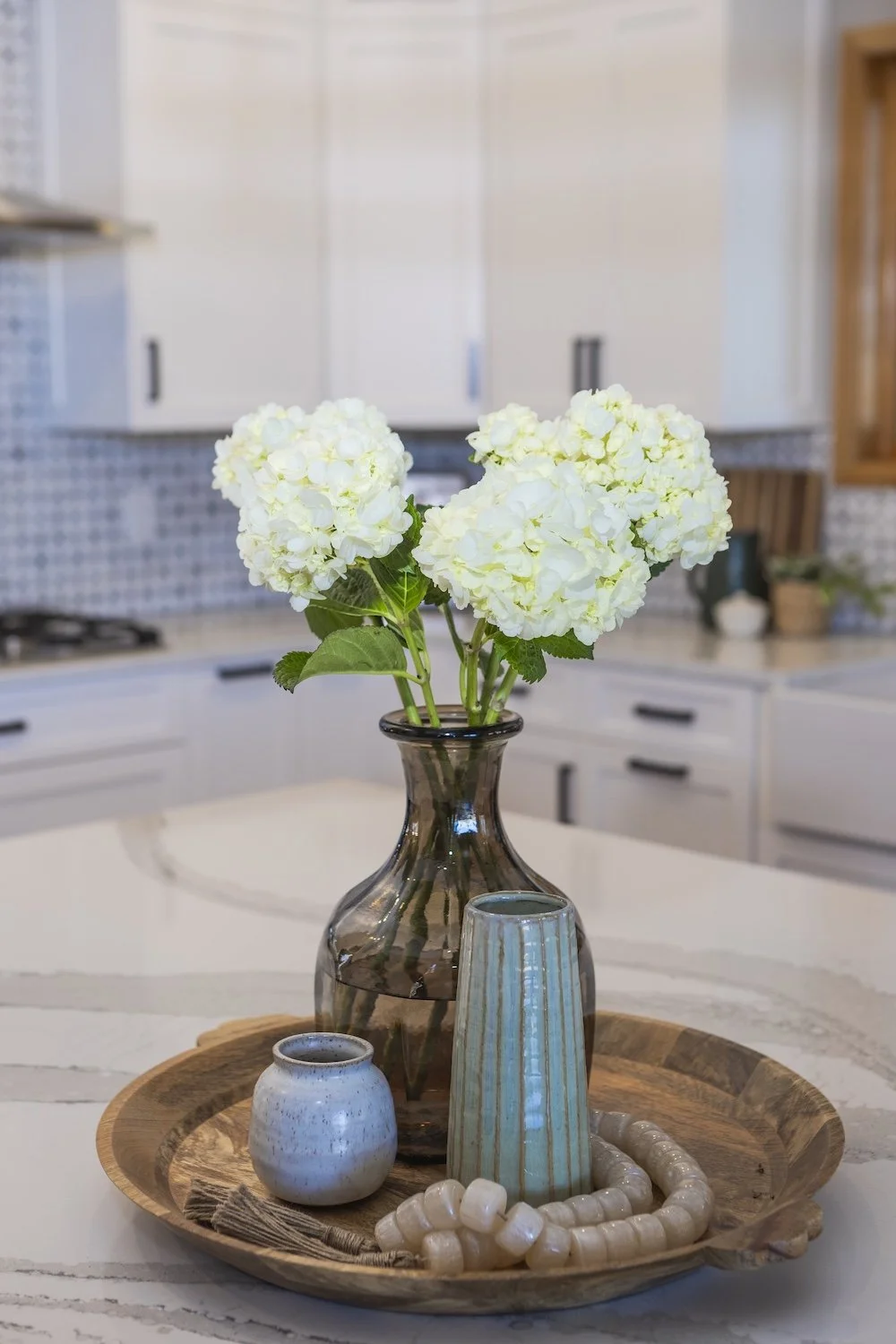 A decorative tray holds a brown glass vase with white hydrangea flowers, a small ceramic pot, a tall striped ceramic vase, a ball of white beads, and a piece of wood on a kitchen countertop.