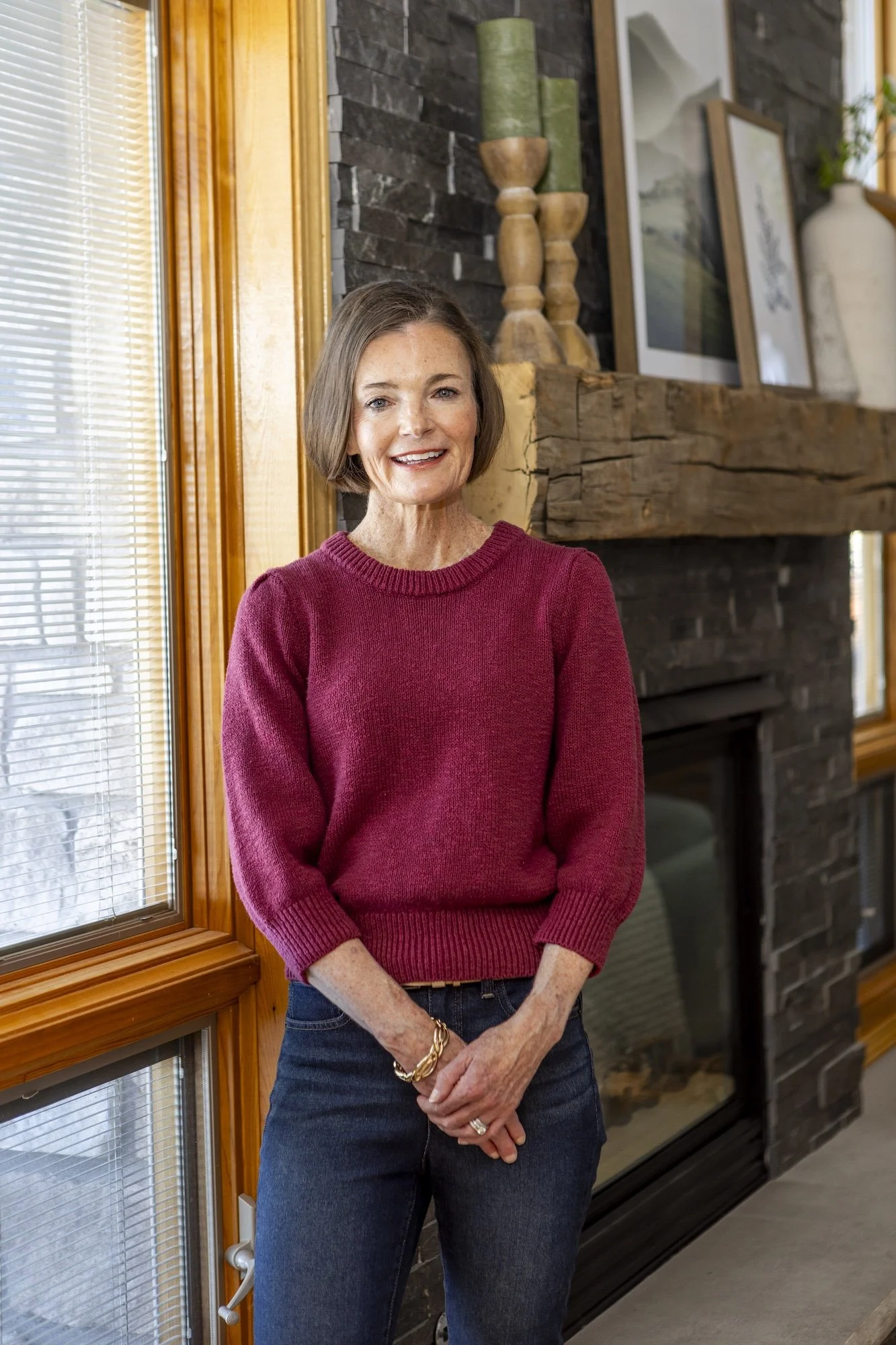 A woman with short brown hair wearing a maroon sweater and dark jeans stands near a large window with blinds. She is smiling and standing inside a room with a stone and wood fireplace and decorated mantle with framed pictures, wooden candle holders, and a large white vase.
