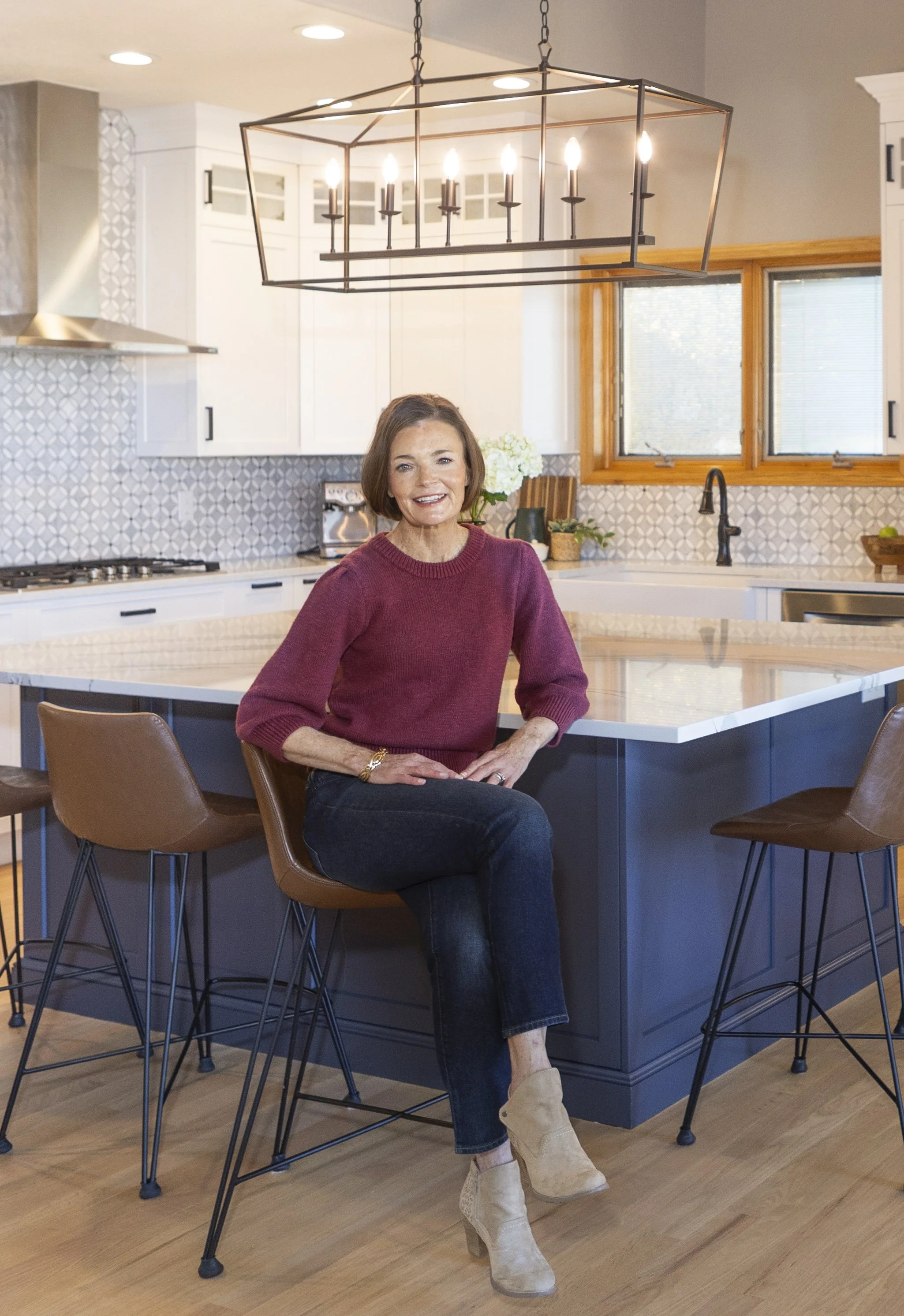 A woman seated on a brown chair in a modern kitchen, smiling, with a large white island, white cabinets, a geometric-patterned backsplash, and a large window in the background.