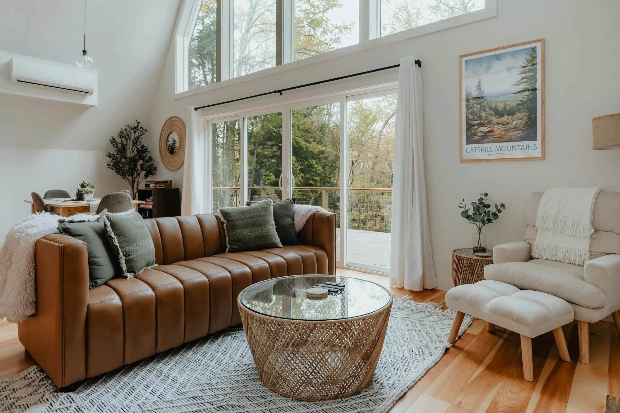 Bright living room with large windows, brown leather sofa with pillows, white armchair with ottoman, glass-top wicker coffee table, and wooden deck visible outside with trees