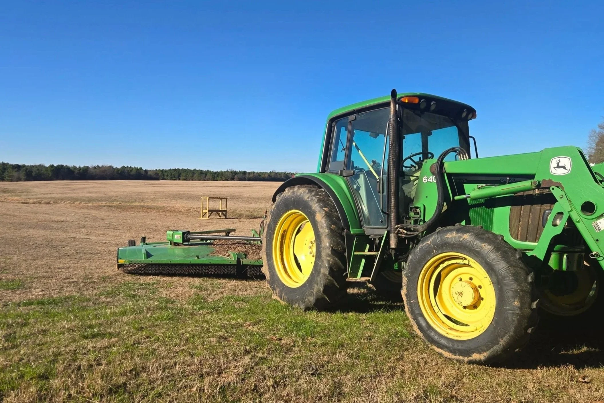 Green John Deere tractor with yellow wheels operating in a field under a clear blue sky, with a bush hog attached.