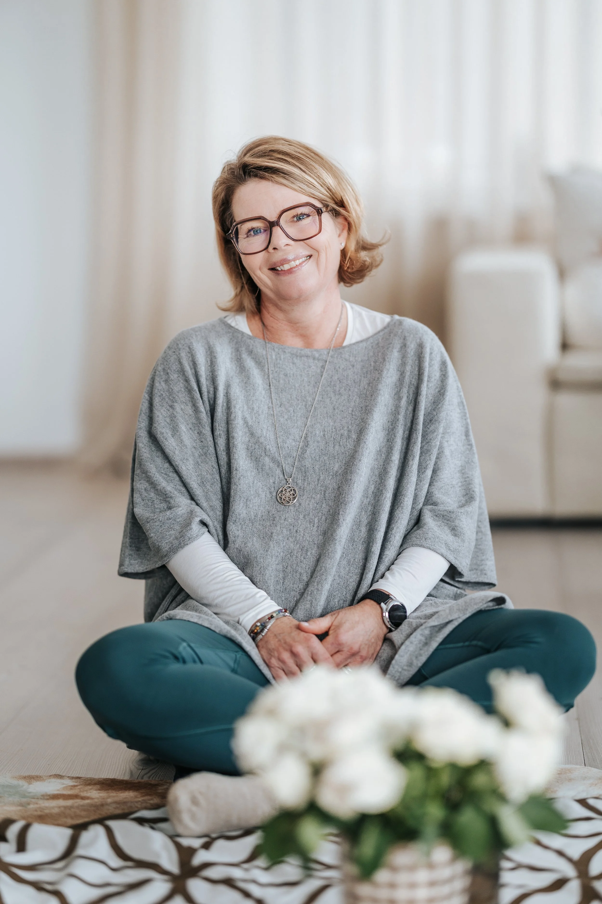A woman sitting cross-legged on a patterned blanket, smiling, with a bouquet of white flowers in front of her in a bright, cozy room.