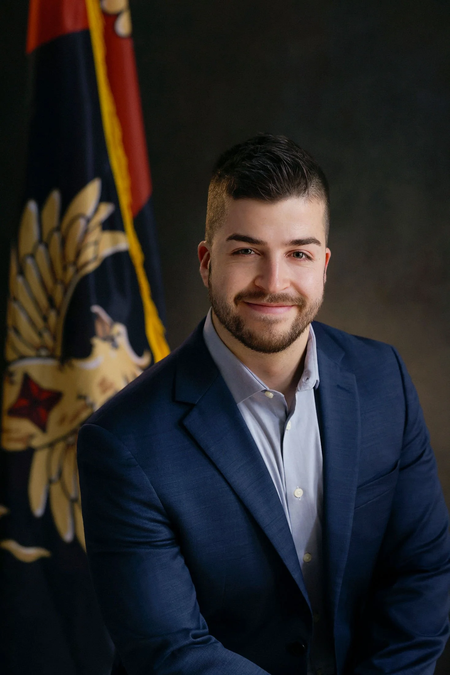 A young man with short dark hair and a beard dressed in a navy blue suit and light blue shirt, sitting in front of a dark background with a flag behind him.
