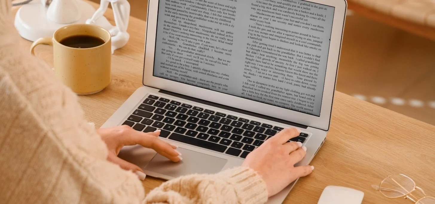 A person wearing a beige sweater typing on a silver laptop with text on the screen, accompanied by a yellow coffee mug, a white teapot, and glasses on a wooden table.