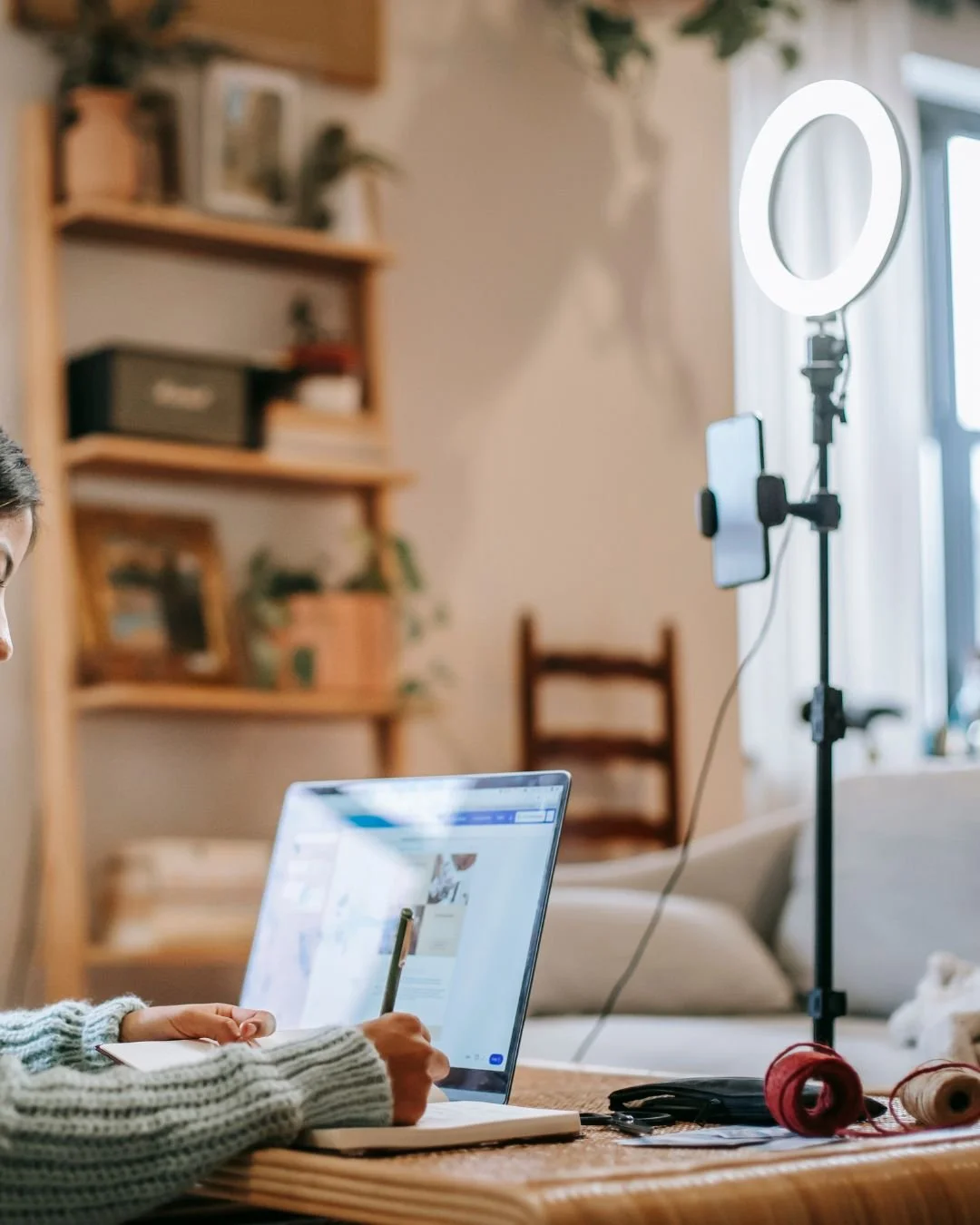Person working on a laptop at a wooden table with a smartphone on a ring light stand and a notebook.