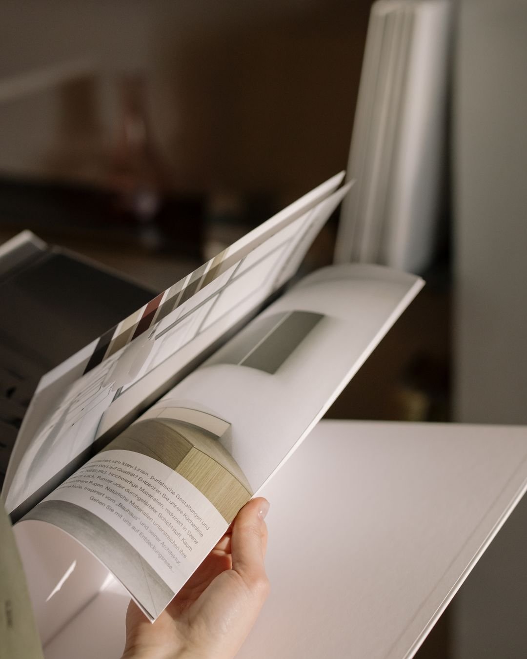 A person holding an open interior design catalog, displaying a page with a modern curved wooden bench or seating arrangement in a neutral color palette.