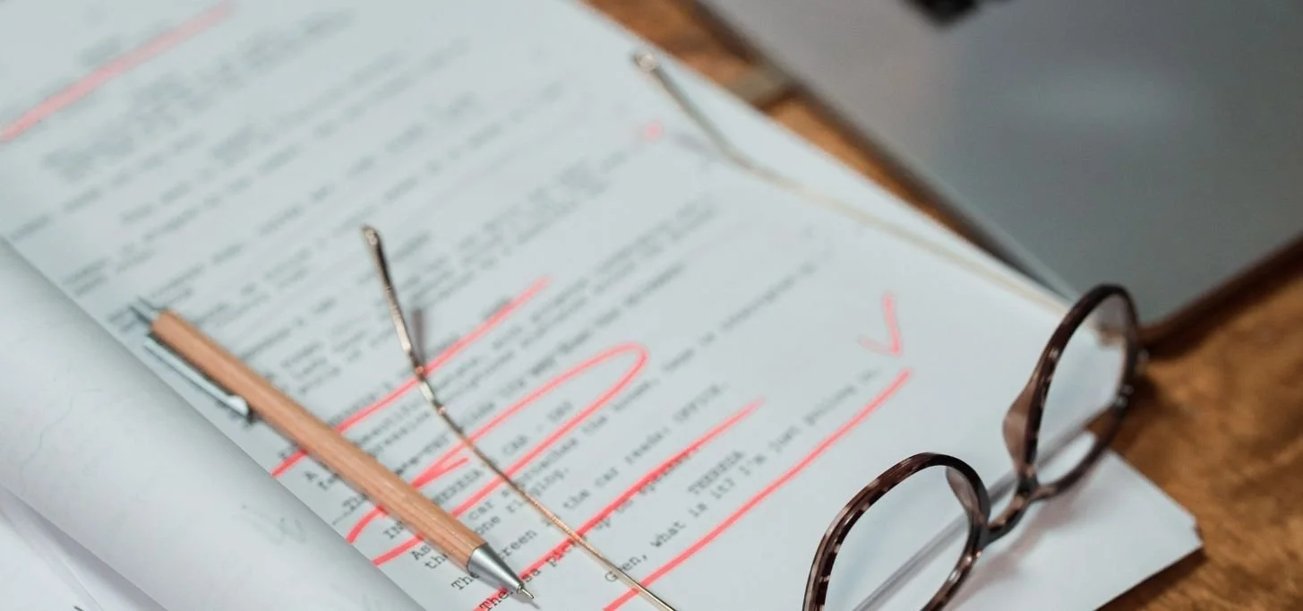Close-up of a manuscript with handwritten edits marked in red ink, a reading glass resting on the page, a pencil, and a book in the background.