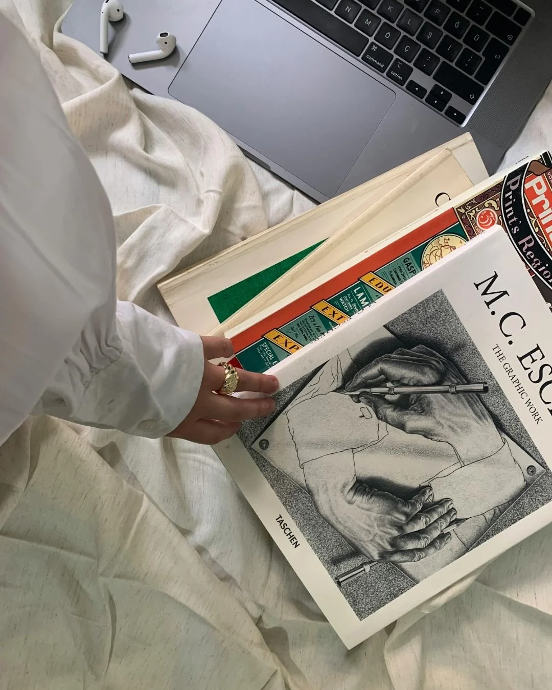 A person holding a large art book titled 'M.C. Escher: The Graphic Work' surrounded by other books, a laptop, and a pair of wireless earbuds on a bed with cream-colored sheets.
