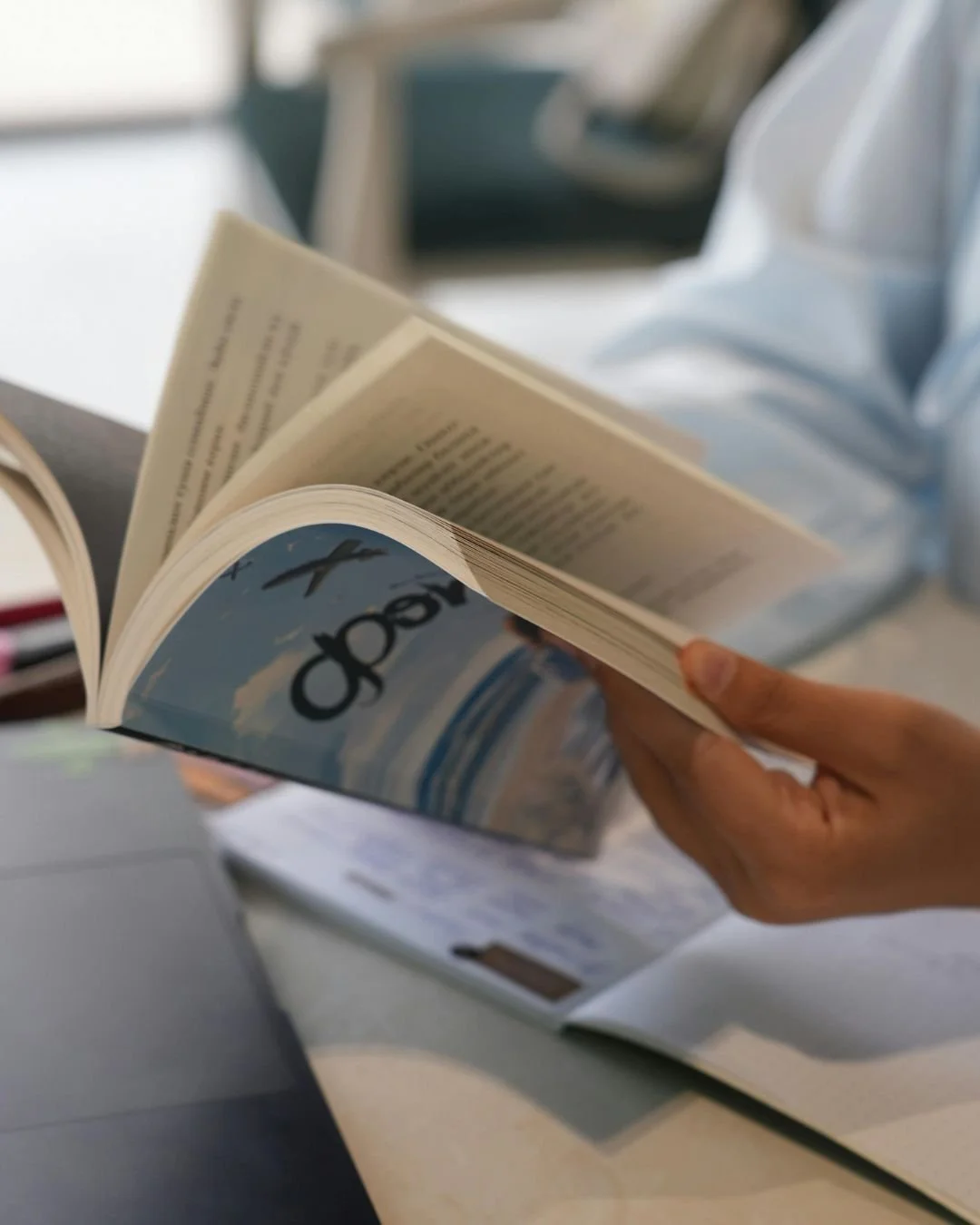A person holding an open book with a beach scene on the cover and the word "Joy" visible, in an indoor setting.