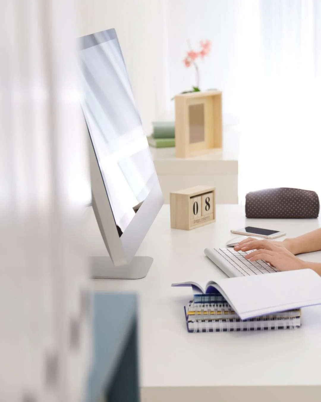 Home office desk with a computer, open notebook, smartphone, and a calendar block displaying 'August 8'. In the background, a flower in a wooden frame.