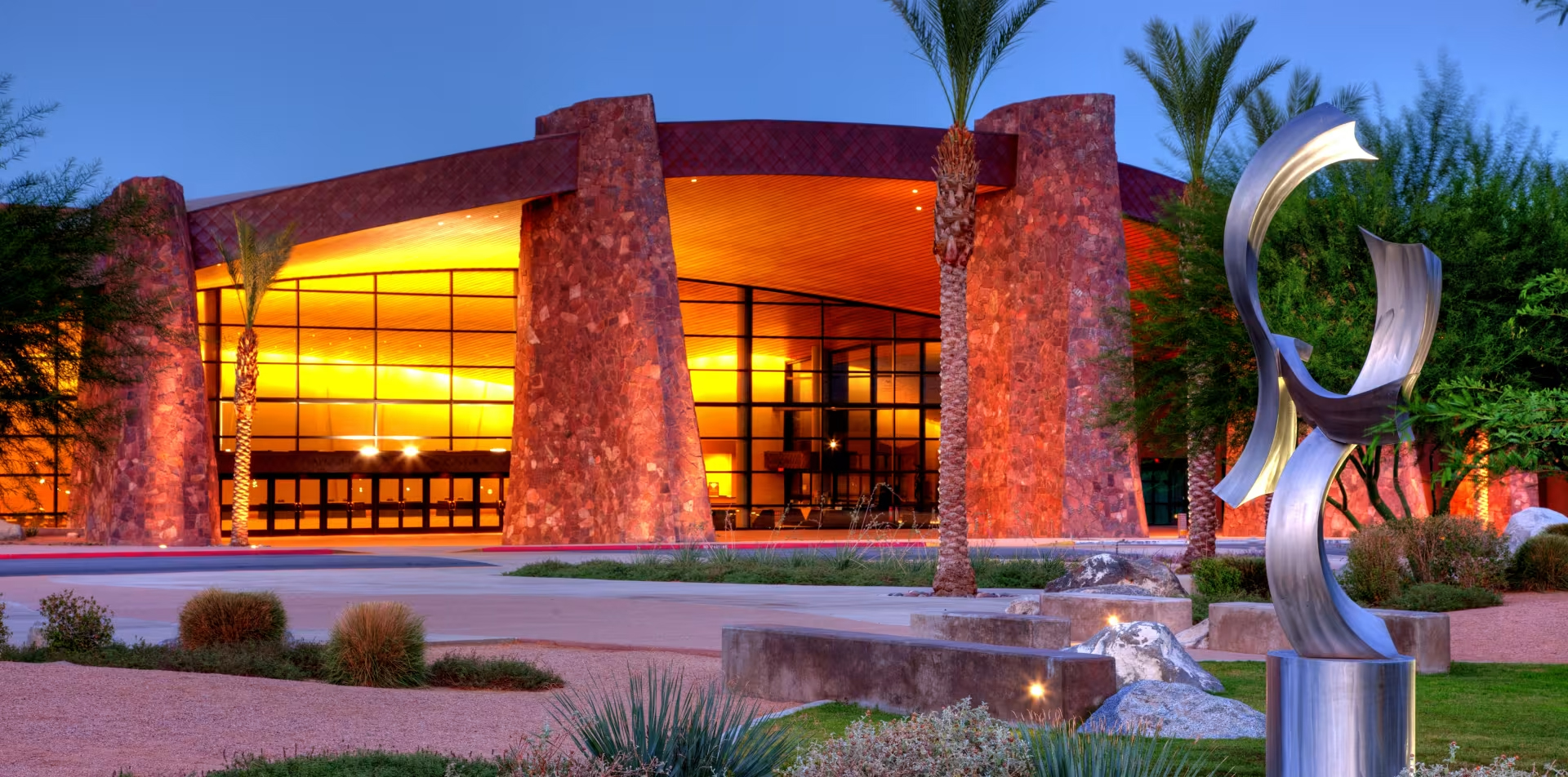 Modern building with a curved roof and large glass windows, illuminated with yellow lighting, surrounded by trees and desert plants, with a metal sculpture in the foreground.