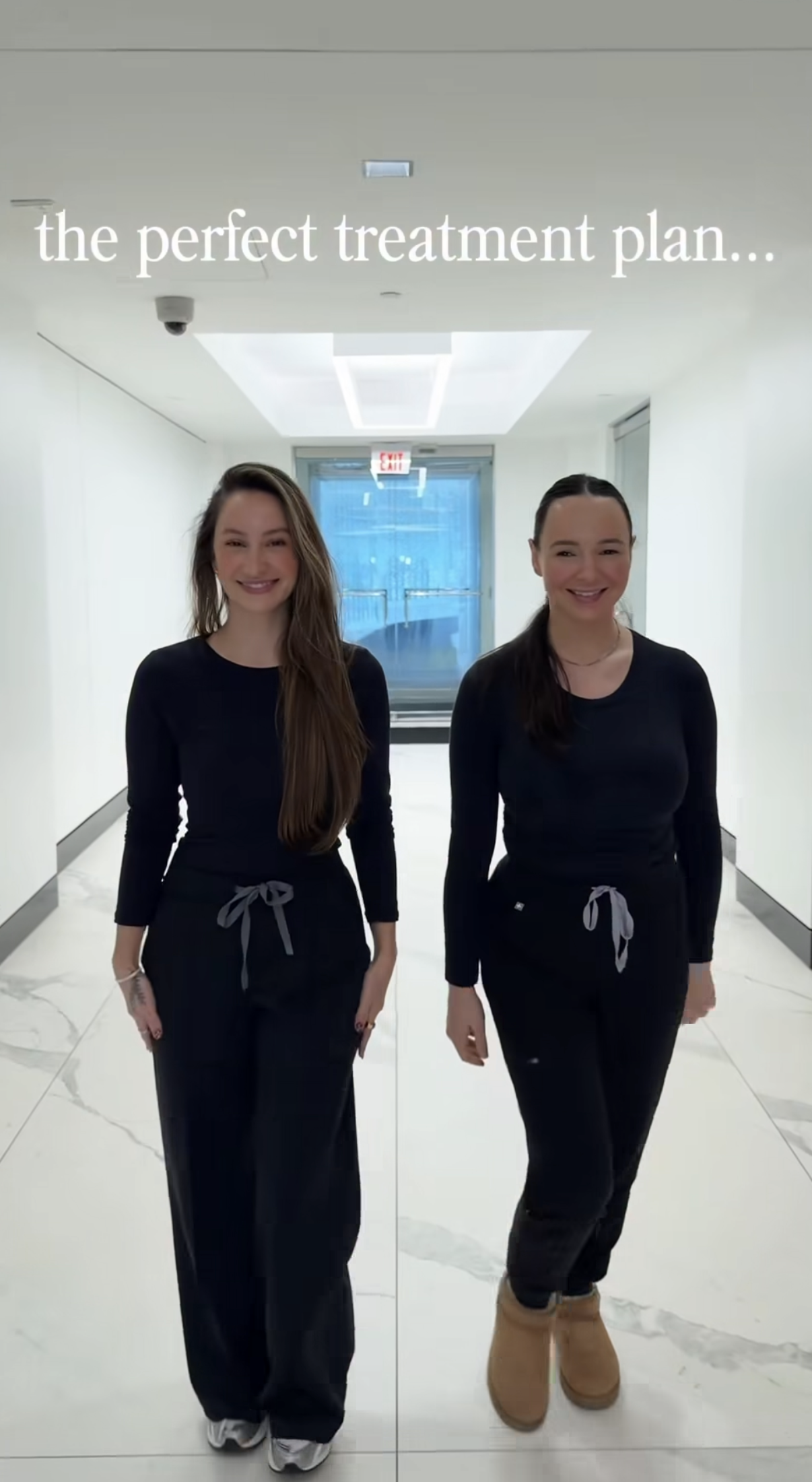 Two women dressed in black standing in a bright hallway with white walls, smiling at the camera, with a sign overhead that reads 'the perfect treatment plan...'
