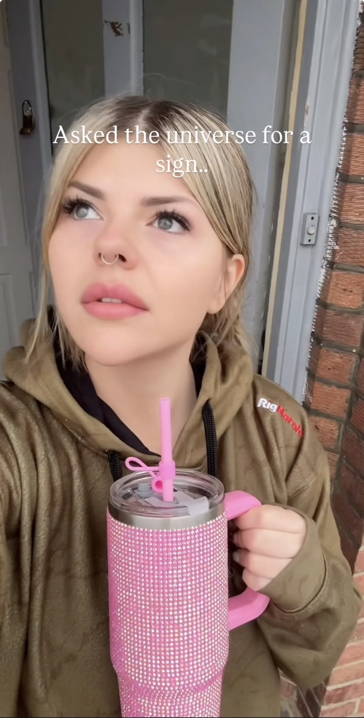 A woman with blonde hair and a septum piercing holding a pink tumbler mug with a straw, standing outside near a brick wall and a door, looking upward with a thought bubble saying 'Asked the universe for a sign..'