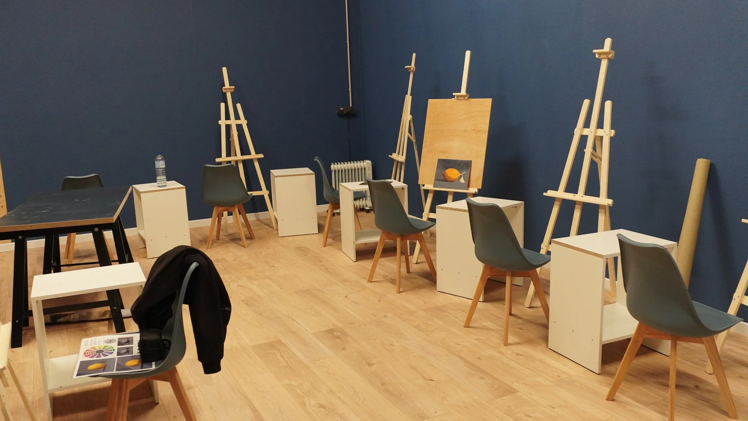 Salle d'art avec plusieurs chevalets, chaises modernes, et petits meubles en bois, murs bleu foncé, sol en parquet, un tableau avec une peinture d'orange, un bouteille d'eau sur une table, et un radiateur