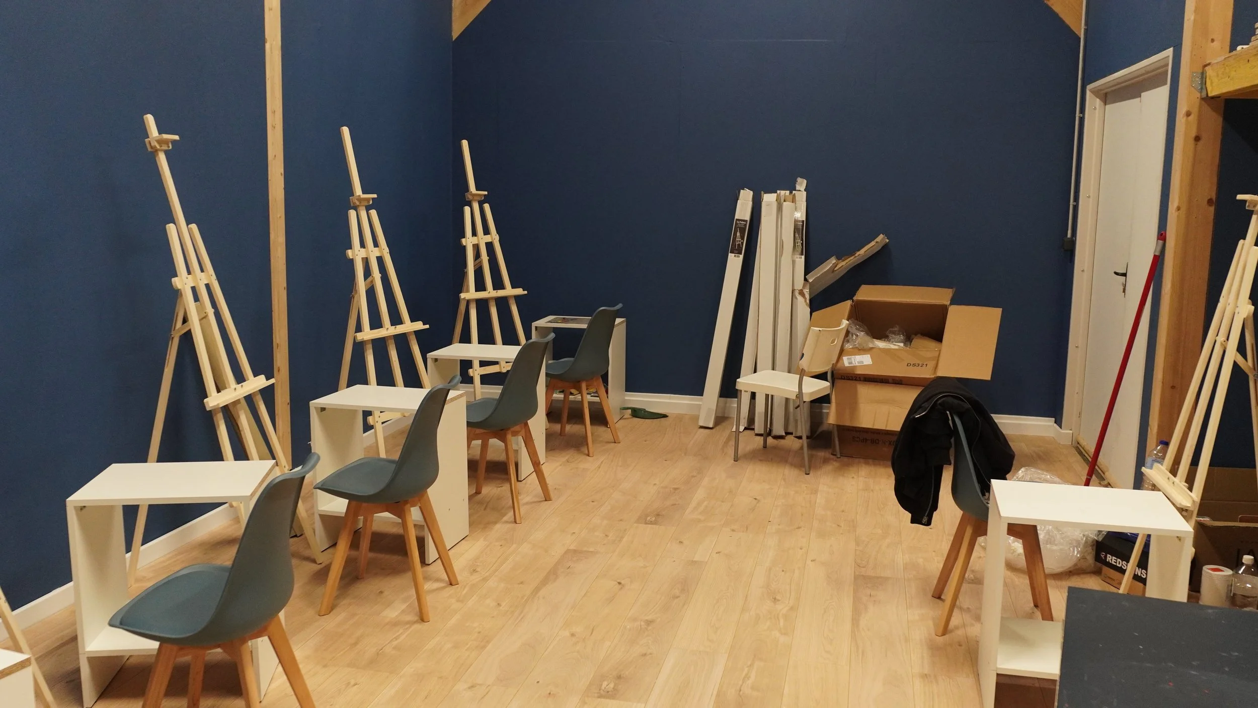 Une pièce en cours d'aménagement avec des chaises, des tables et des supports pour tableaux, quelques boîtes en carton, un balai et des matériaux de construction. Le mur est peint en bleu.