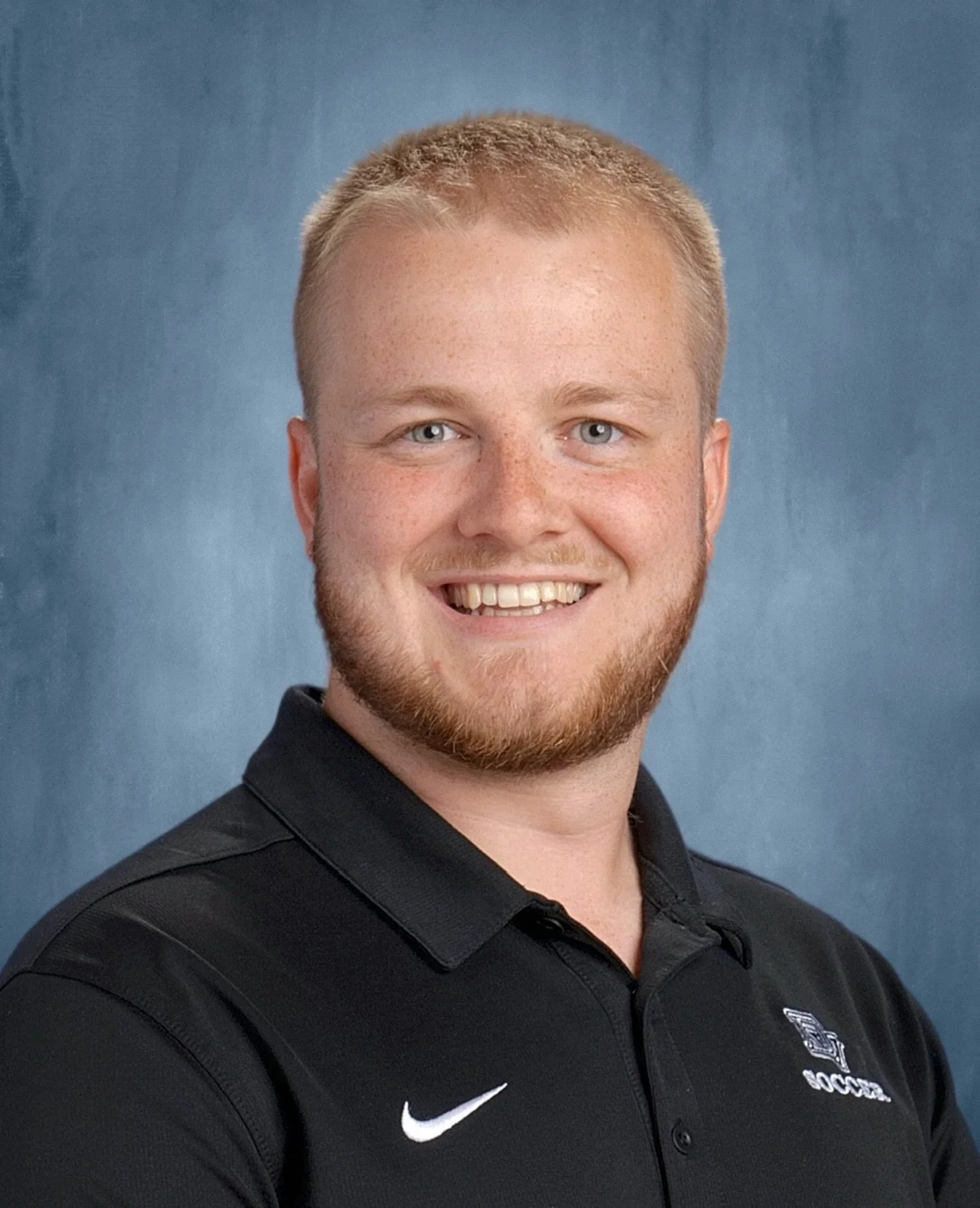 Headshot of a smiling young man with short blond hair, facial hair, wearing a black polo shirt with a Nike logo and a school logo, against a blue background.