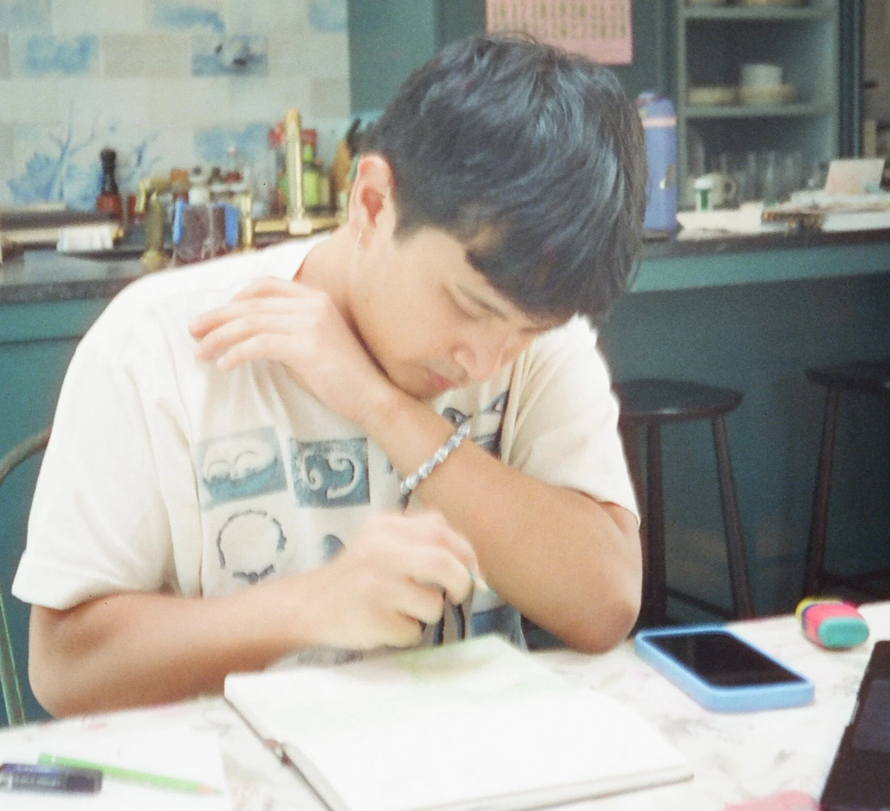 A young boy with black hair and light skin sitting at a table in a classroom, engaging with a mobile device, with school supplies on the table and classroom decor visible in the background.
